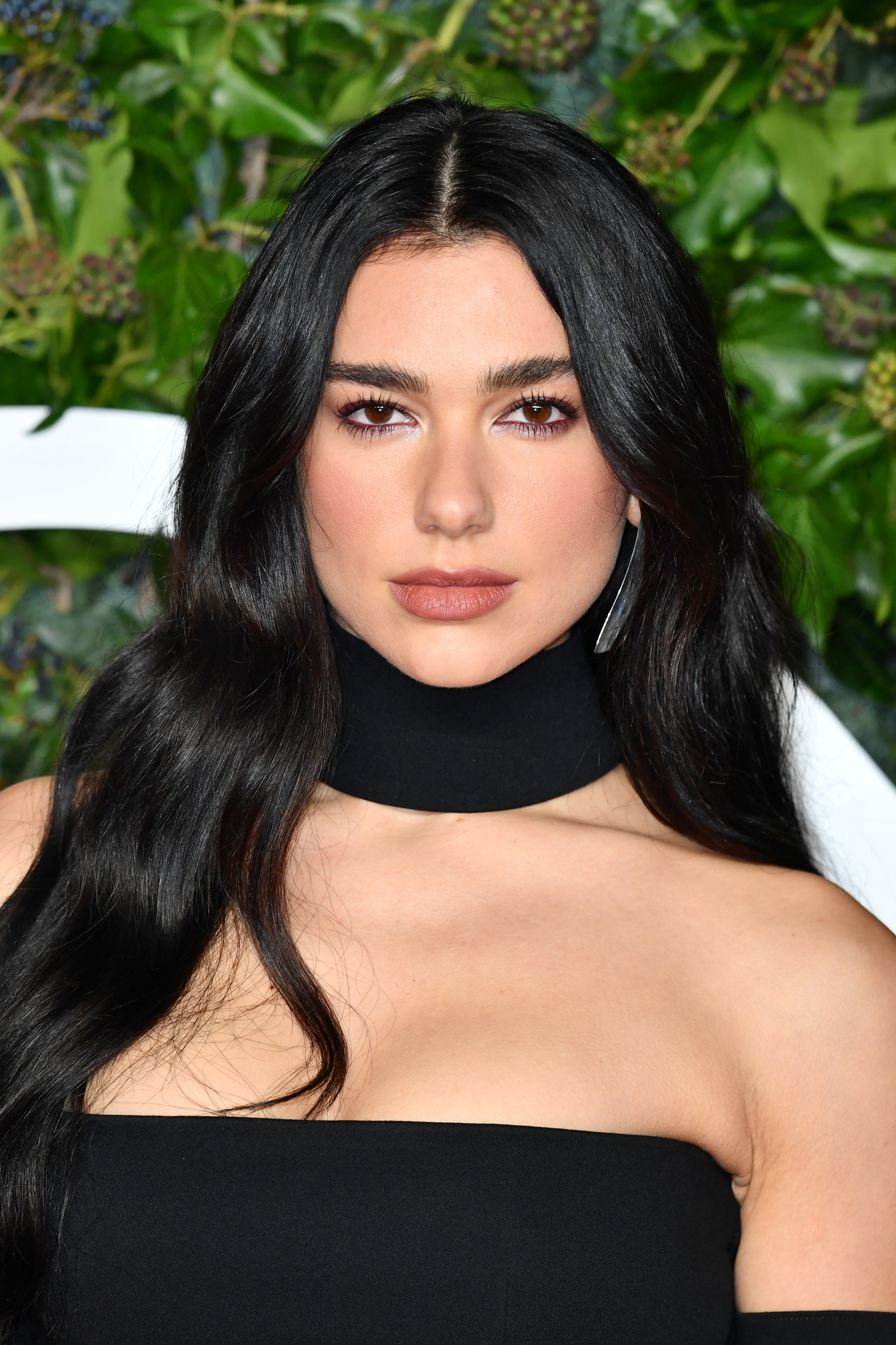 Person with long black hair wearing an off-the-shoulder black outfit, standing in front of a leafy backdrop