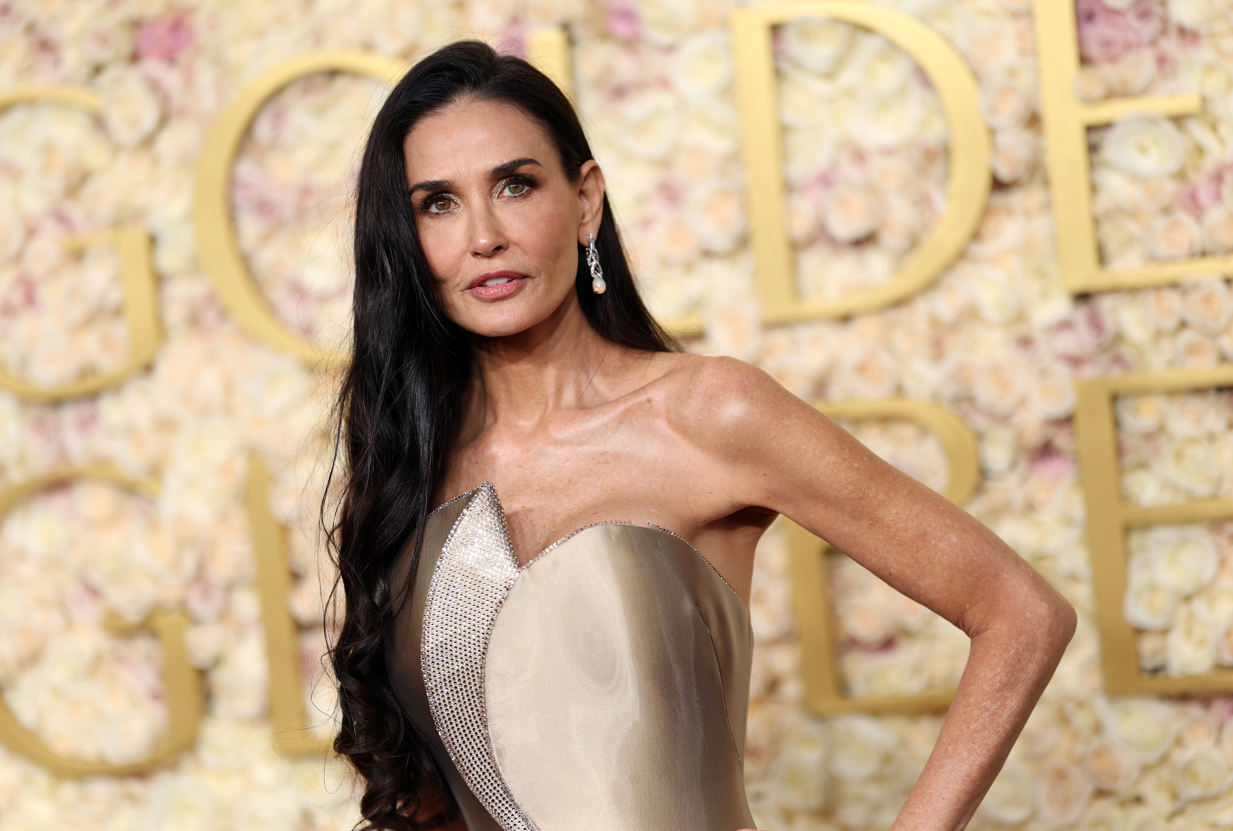 Demi Moore in an elegant strapless gown with textured detailing poses at an event, standing in front of a flower and gold-themed backdrop