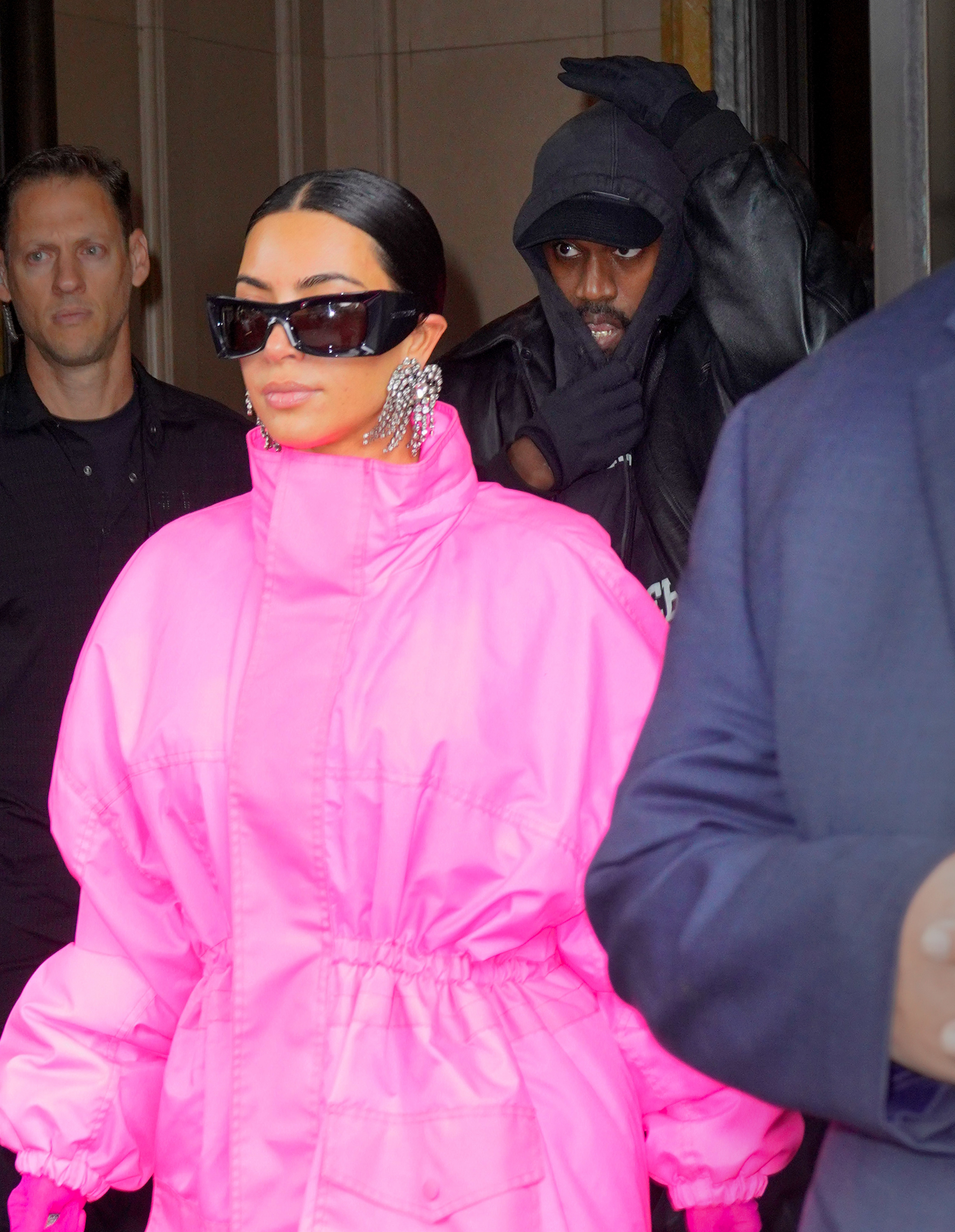 Kim in bright, stylish outerwear and large earrings outside a building, accompanied by Kanye in a hooded jacket and sunglasses