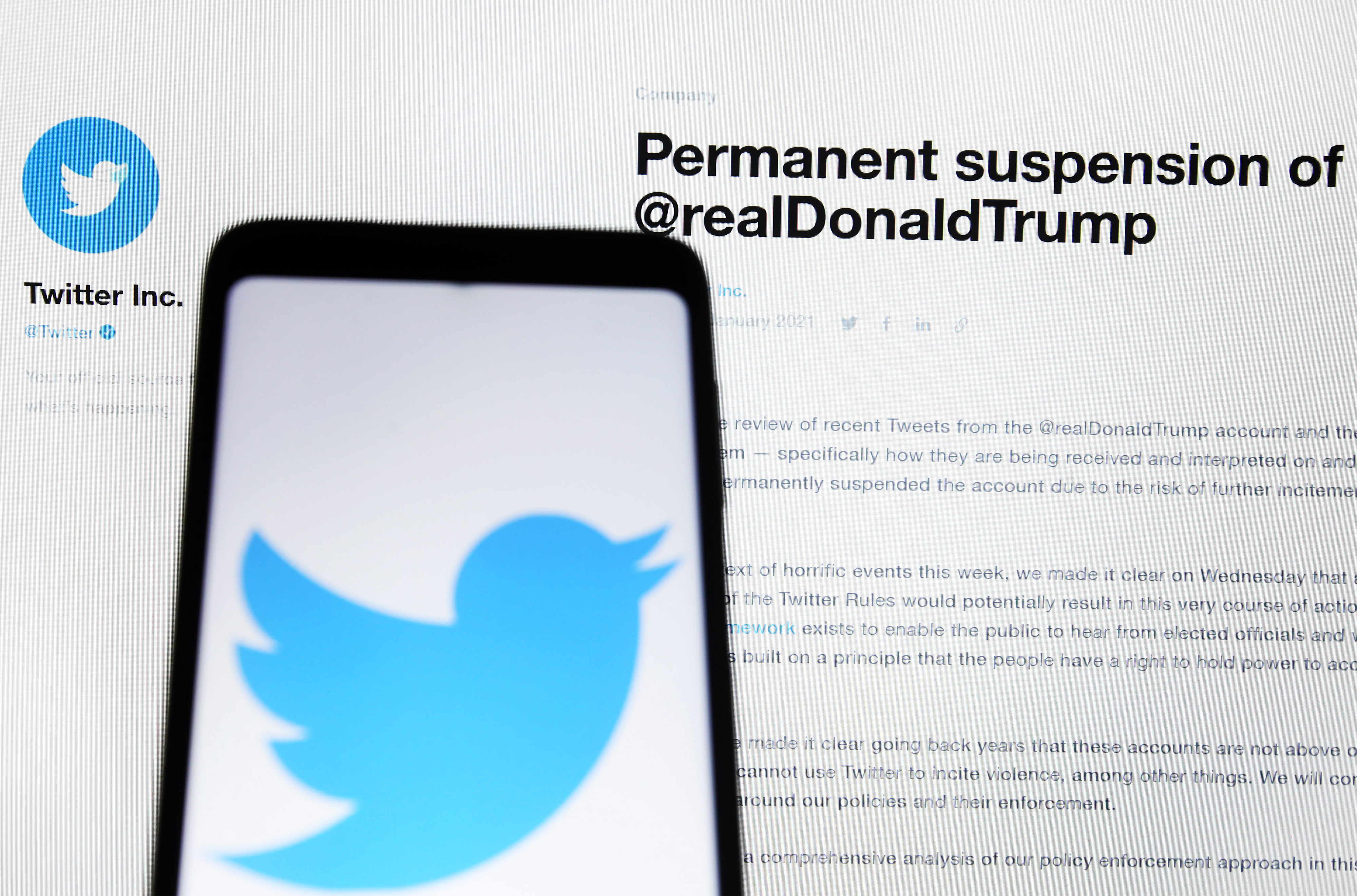 Phone displaying Twitter logo with background text about the permanent suspension of Trump&#x27;s account on Twitter