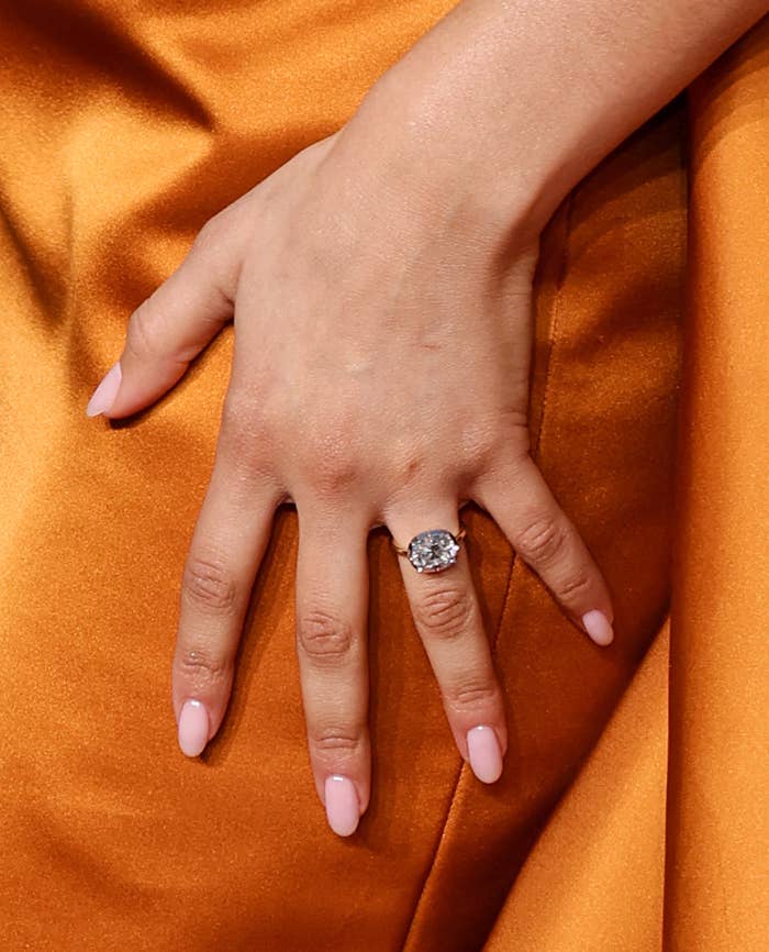 A manicured hand with light pink nails rests on a fabric, showcasing a large, elegant diamond ring