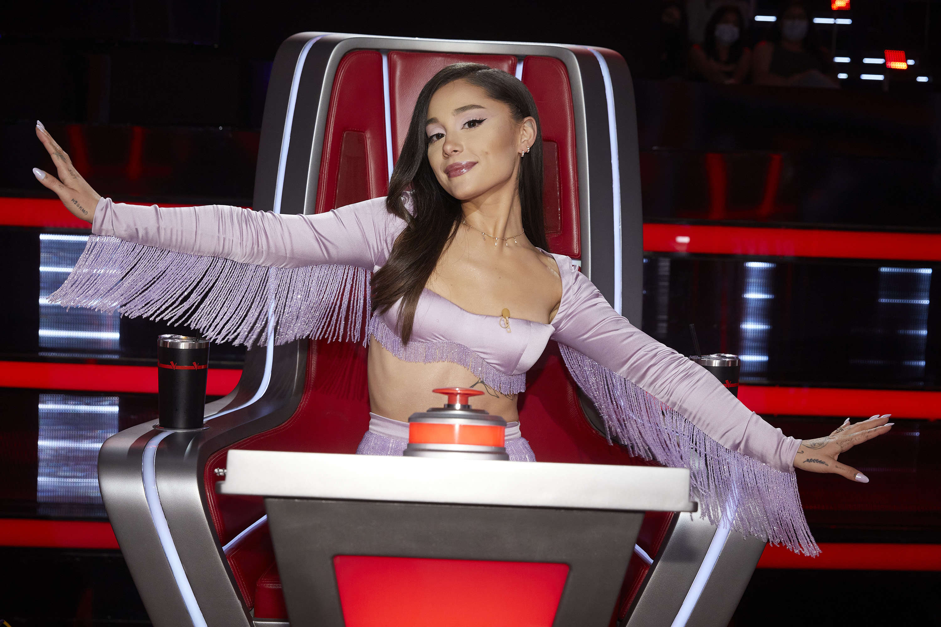 Person posing on a TV show set in a fringe outfit, sitting in a red and silver chair with a button