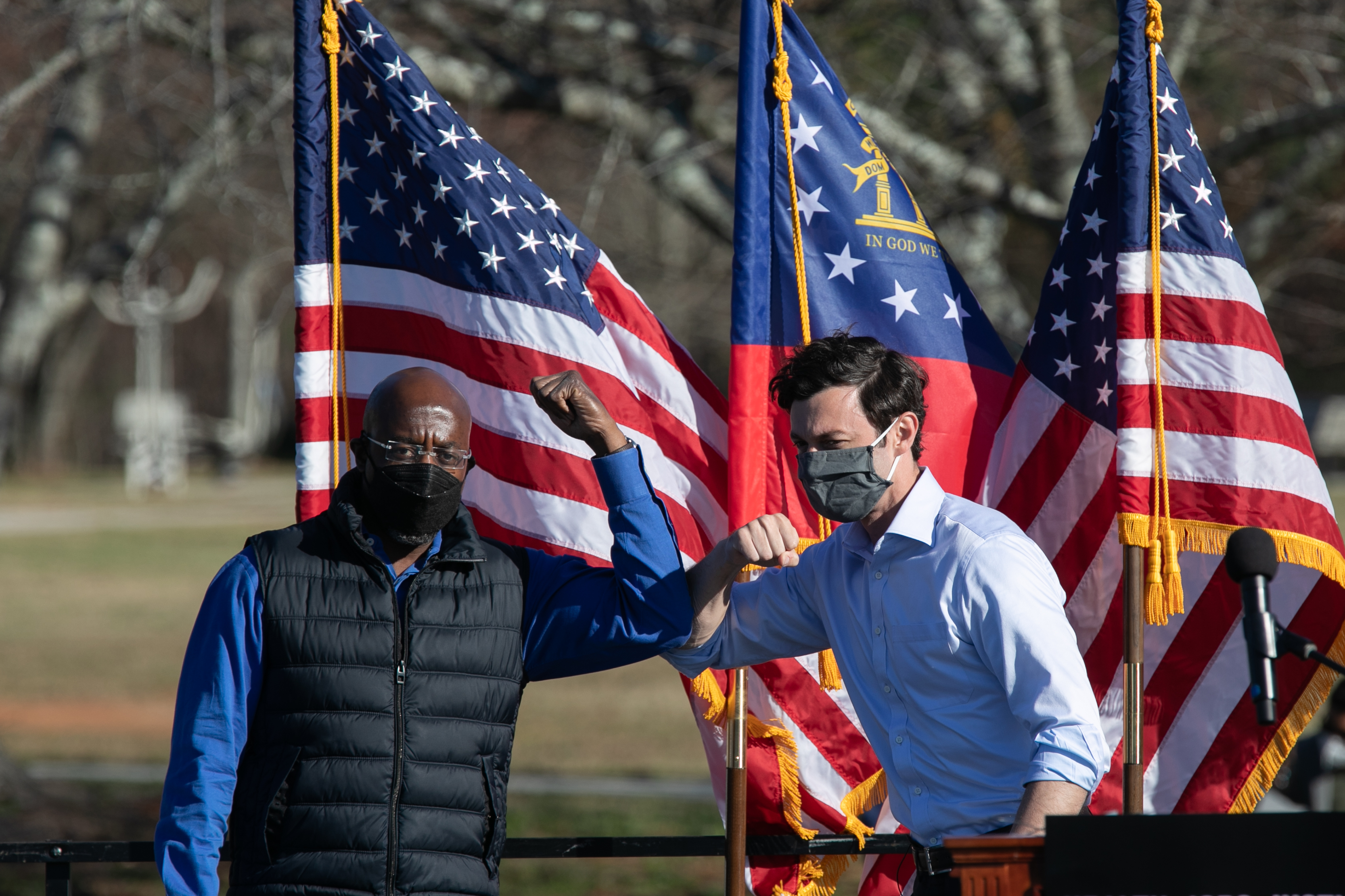 Two people in masks do an elbow bump in front of two American flags during an outdoor event. One wears a vest and the other a dress shirt