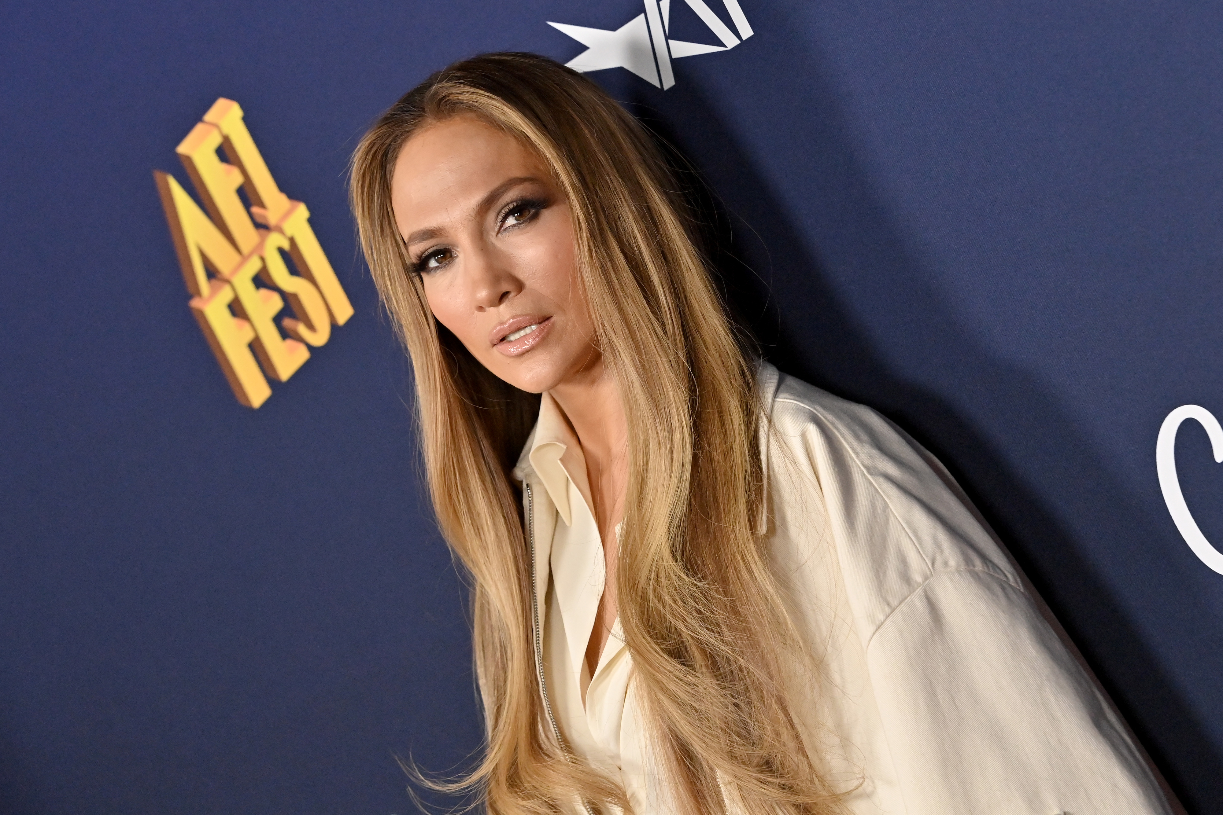 J.Lo with long hair in a flowing outfit poses at a formal event against a branded backdrop