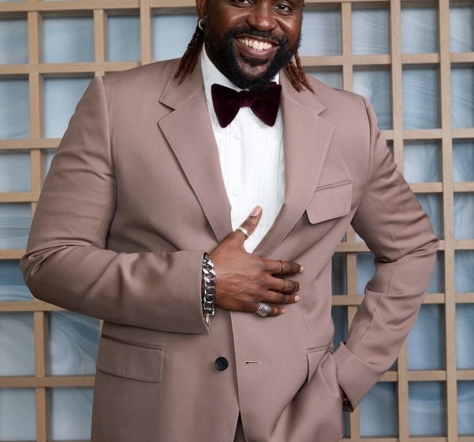 Brian Tyree Henry in a tailored suit with a bow tie, standing and smiling in front of a wooden grid backdrop