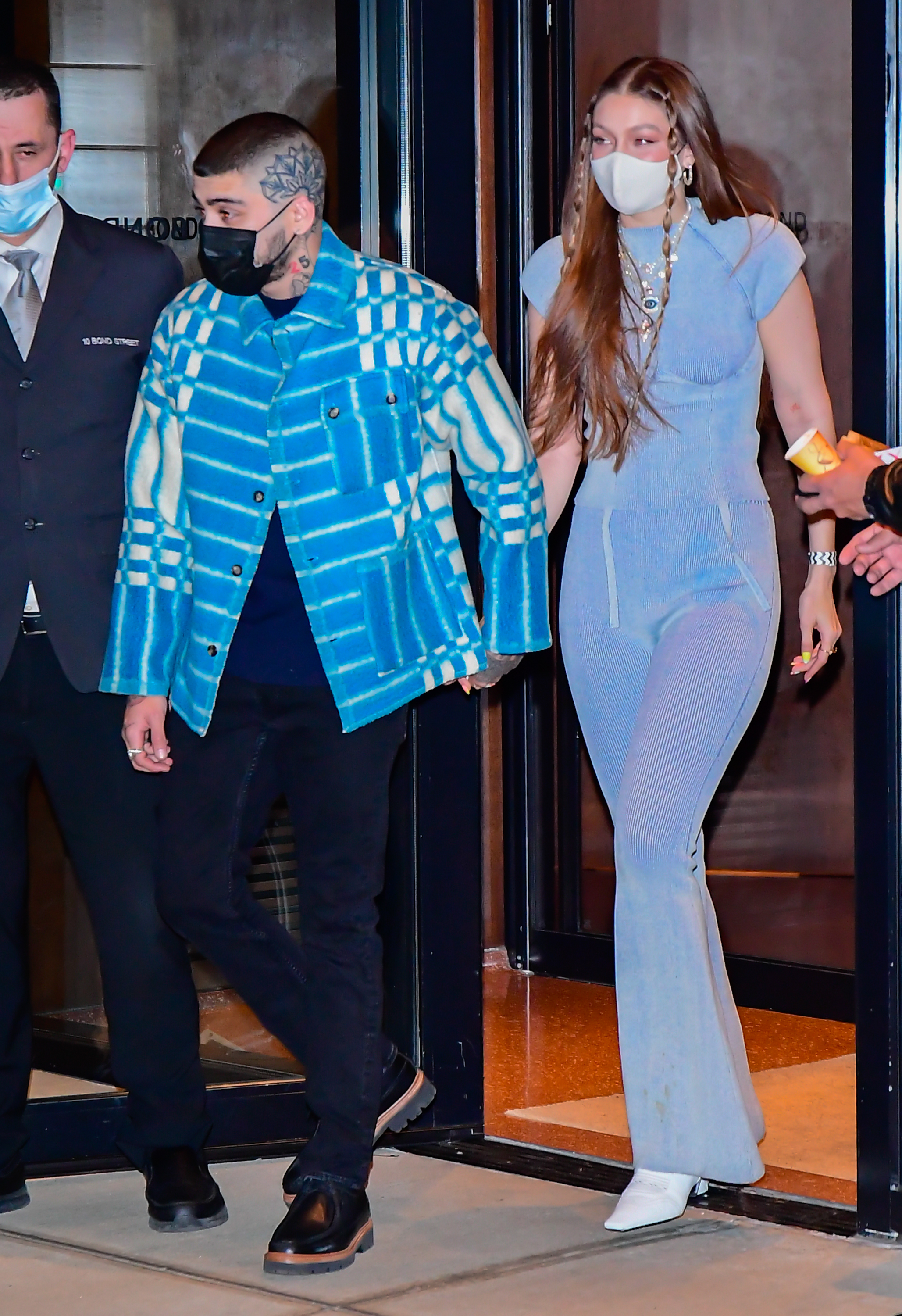 A man in a patterned jacket and a woman in a fitted jumpsuit, both wearing masks, walk on a red carpet with security