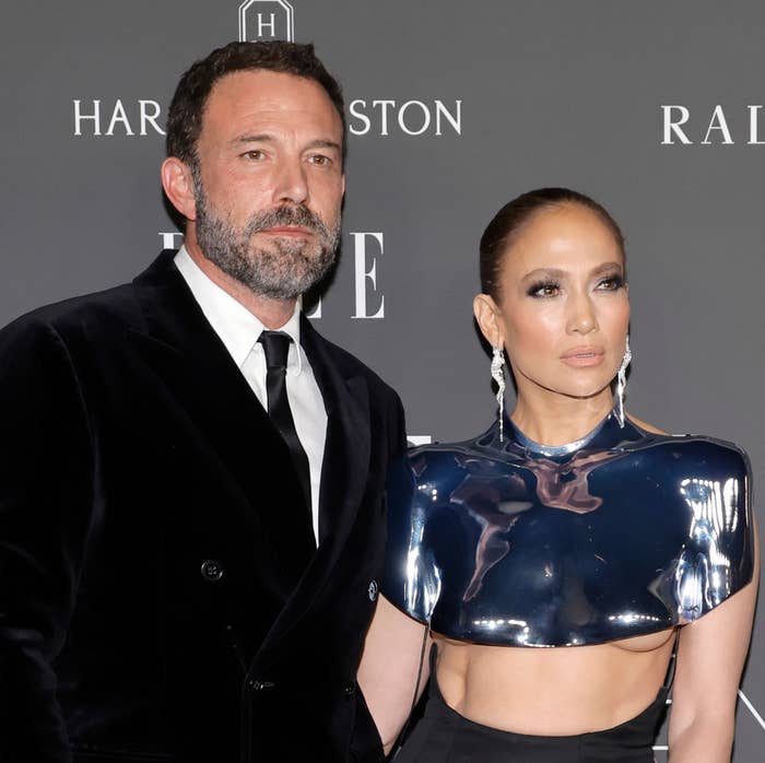 Ben Affleck and J.Lo on a red carpet; one in a black suit and tie, the other in a metallic, futuristic top with cut-outs