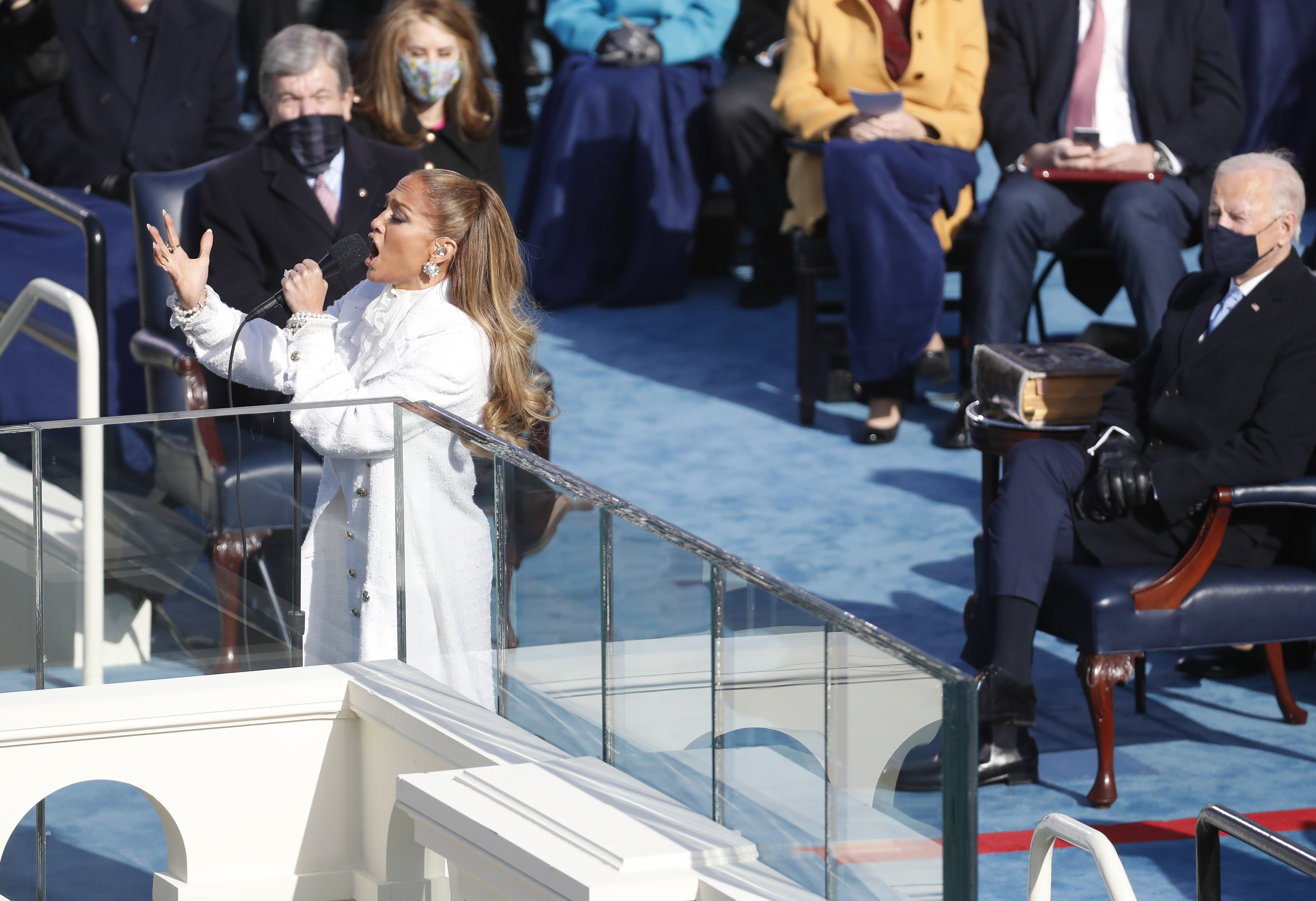 A performer in a white outfit sings at an outdoor event. Distinguished guests are seated behind her on a stage