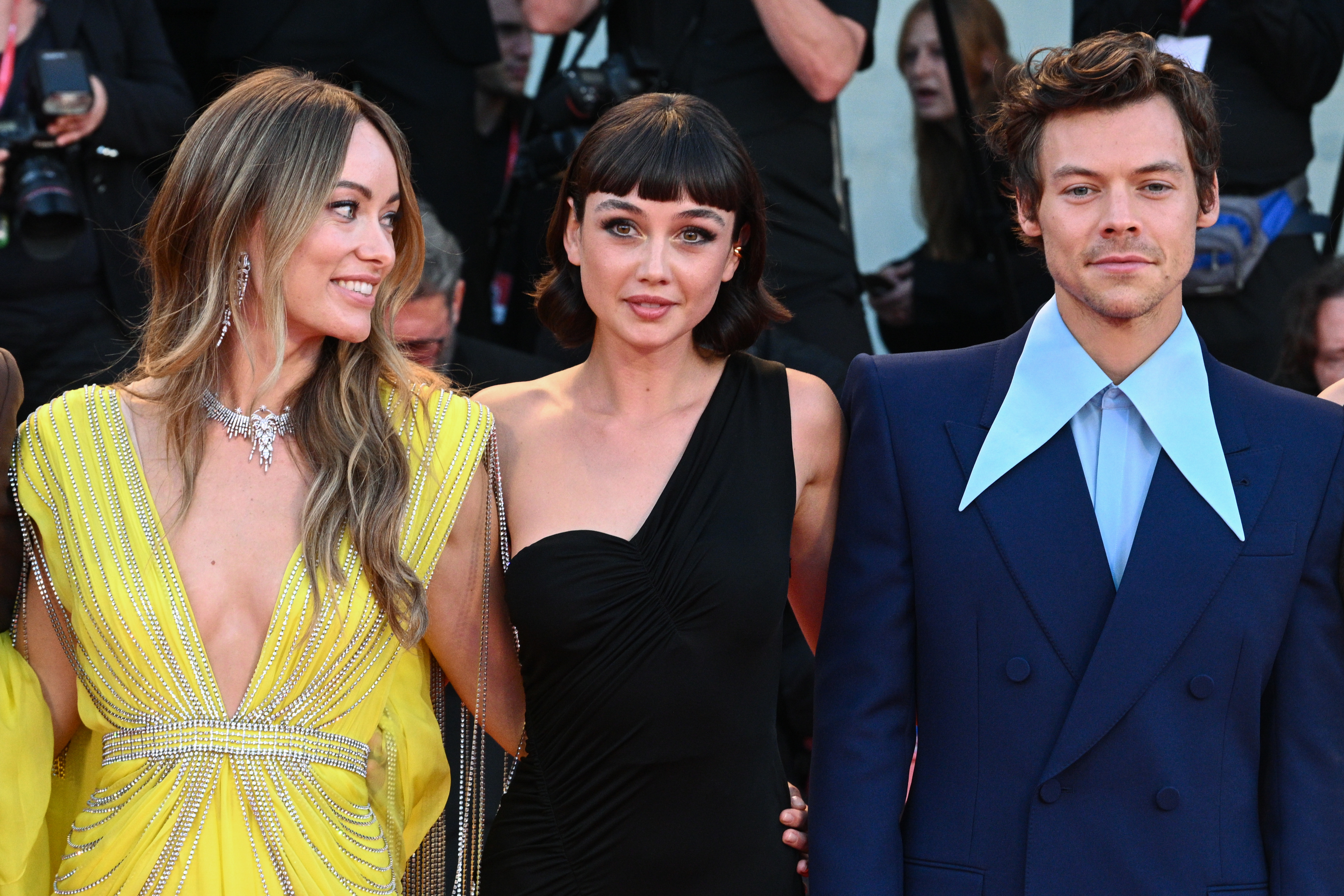 Three people pose on the red carpet. The woman on the left wears an elegant gown, the woman in the middle wears a black dress, and the man wears a suit