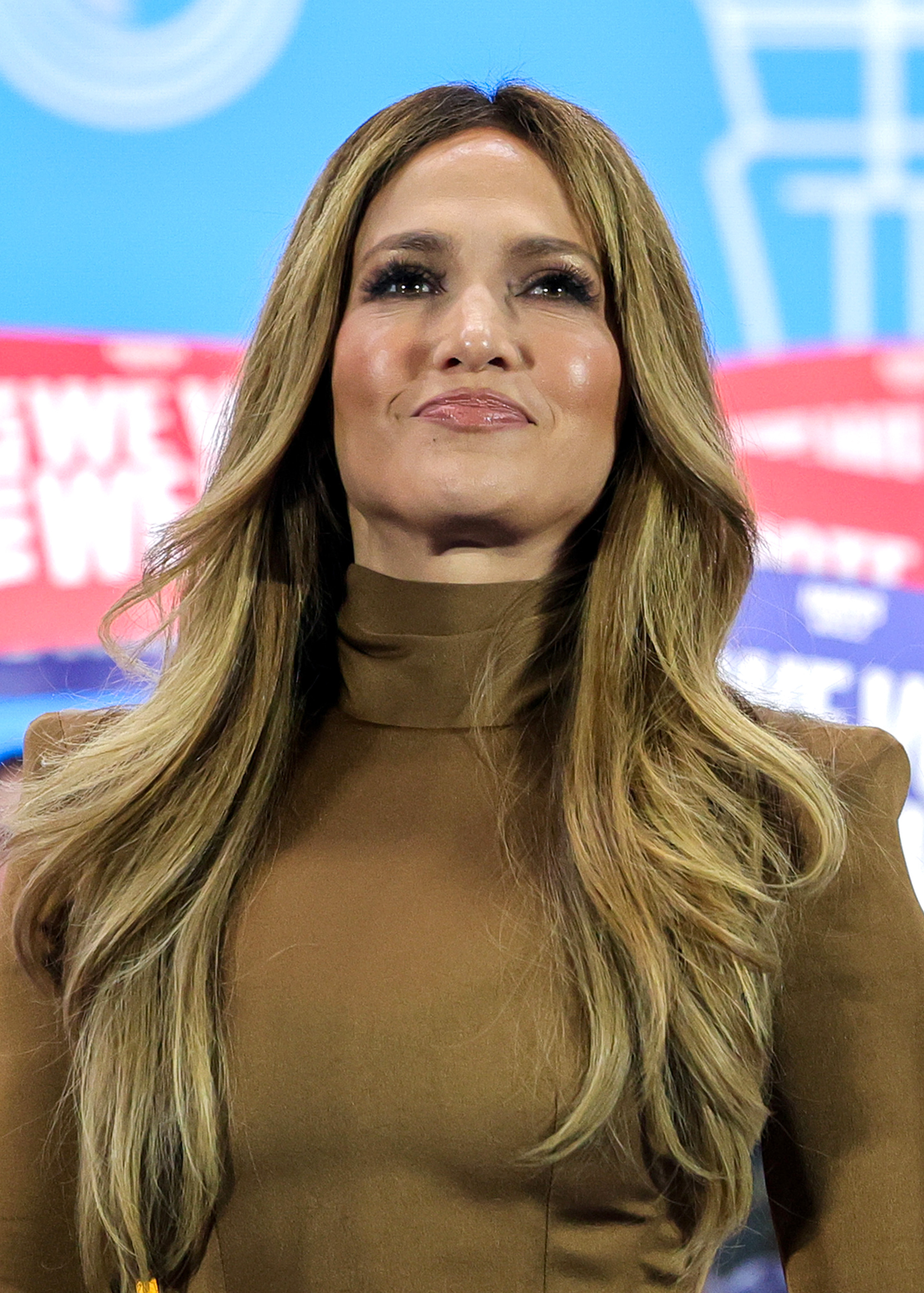 J.Lo with long hair in a high-neck, fitted long-sleeve dress, standing in front of a backdrop with blurred text and graphics