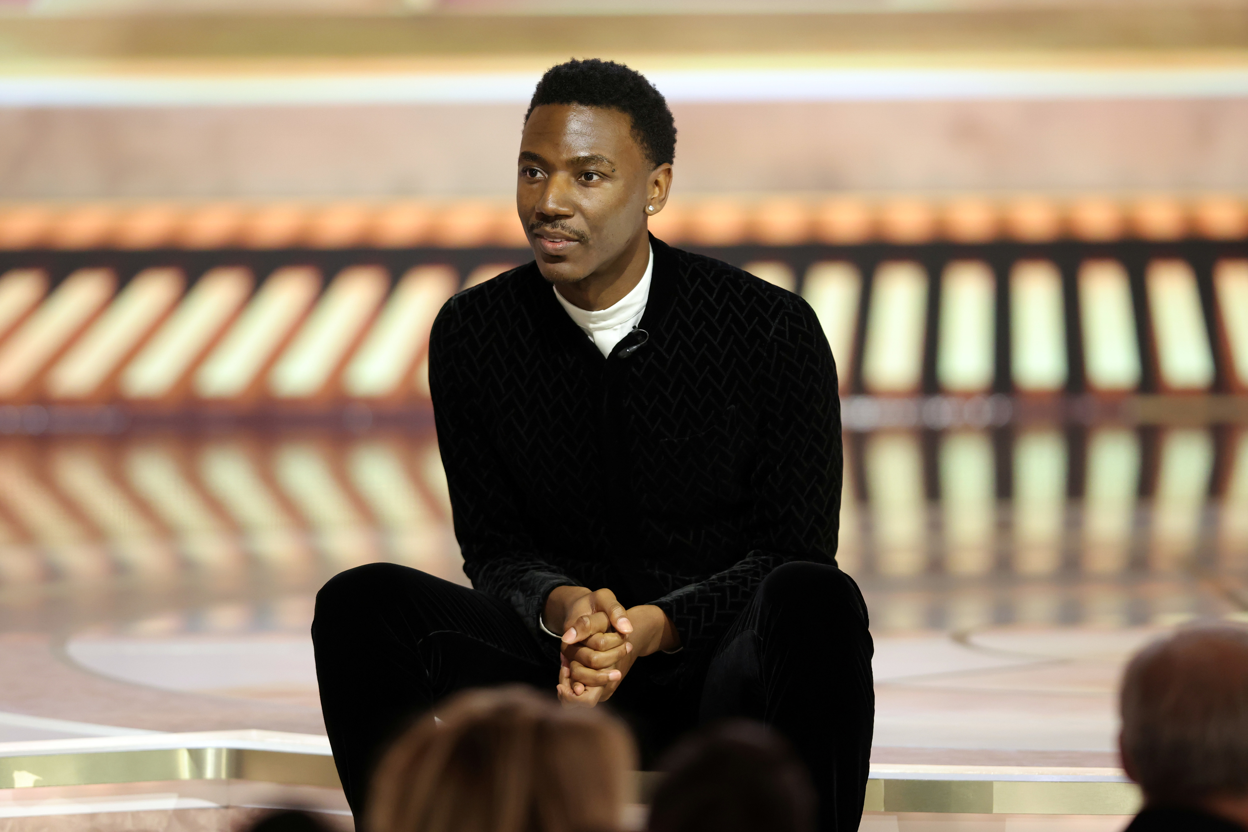 Person seated on stage, wearing a dark textured suit and white shirt, looking calm and composed during an event