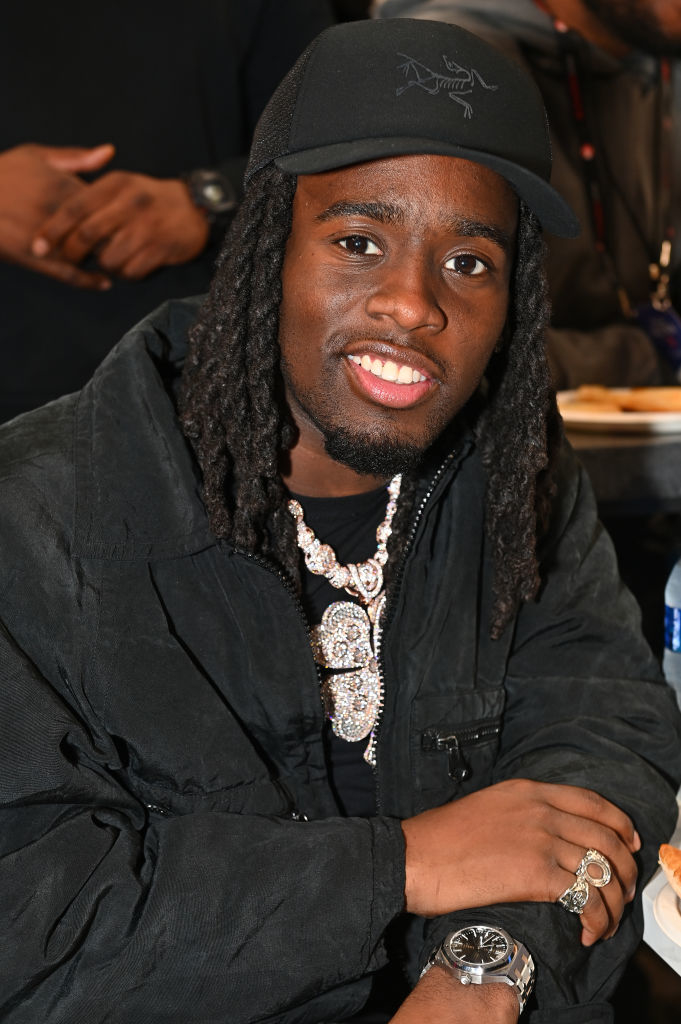 Kai Cenat smiling, wearing a cap, a dark jacket, and layered sparkling necklaces, seated indoors at an event