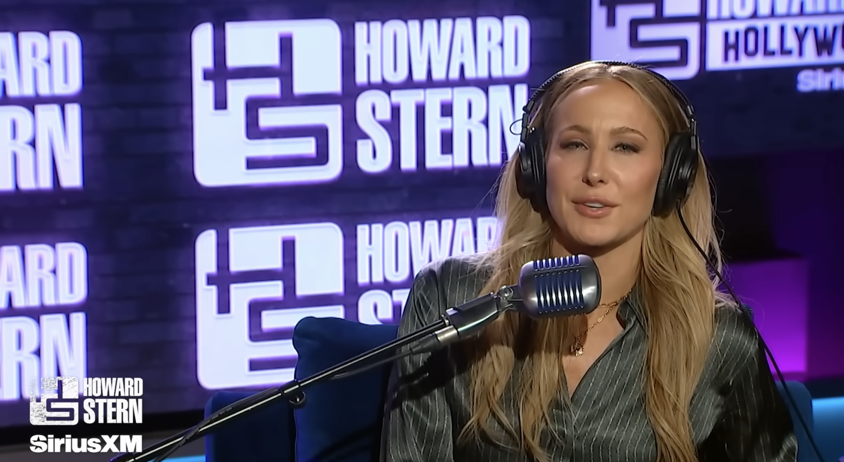 Person in a studio wearing headphones and speaking into a microphone, with &quot;Howard Stern&quot; and &quot;SiriusXM&quot; logos visible in the background