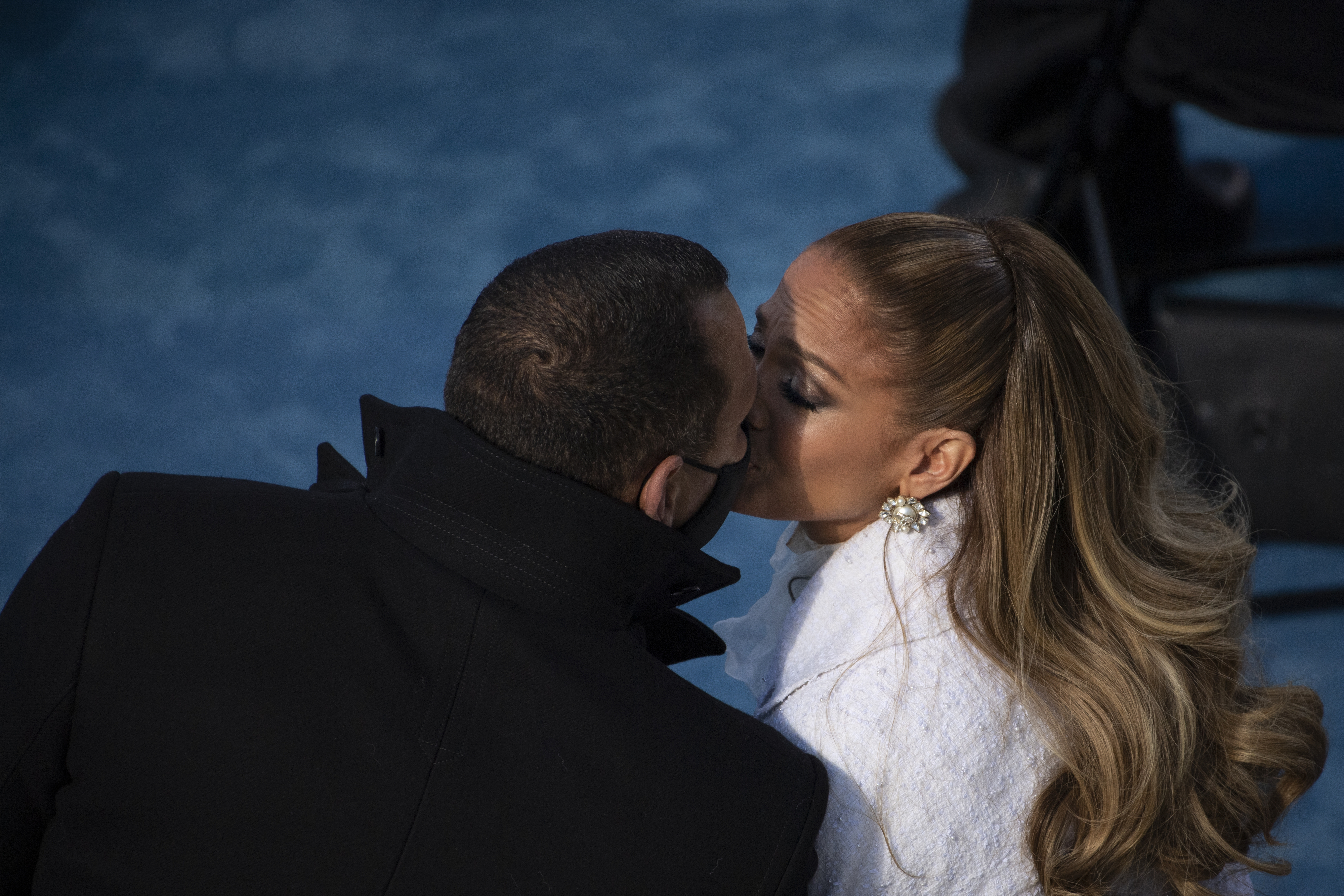 Two people, in elegant attire, share a kiss. The woman has a fashionable ponytail and wears large earrings. The event seems formal