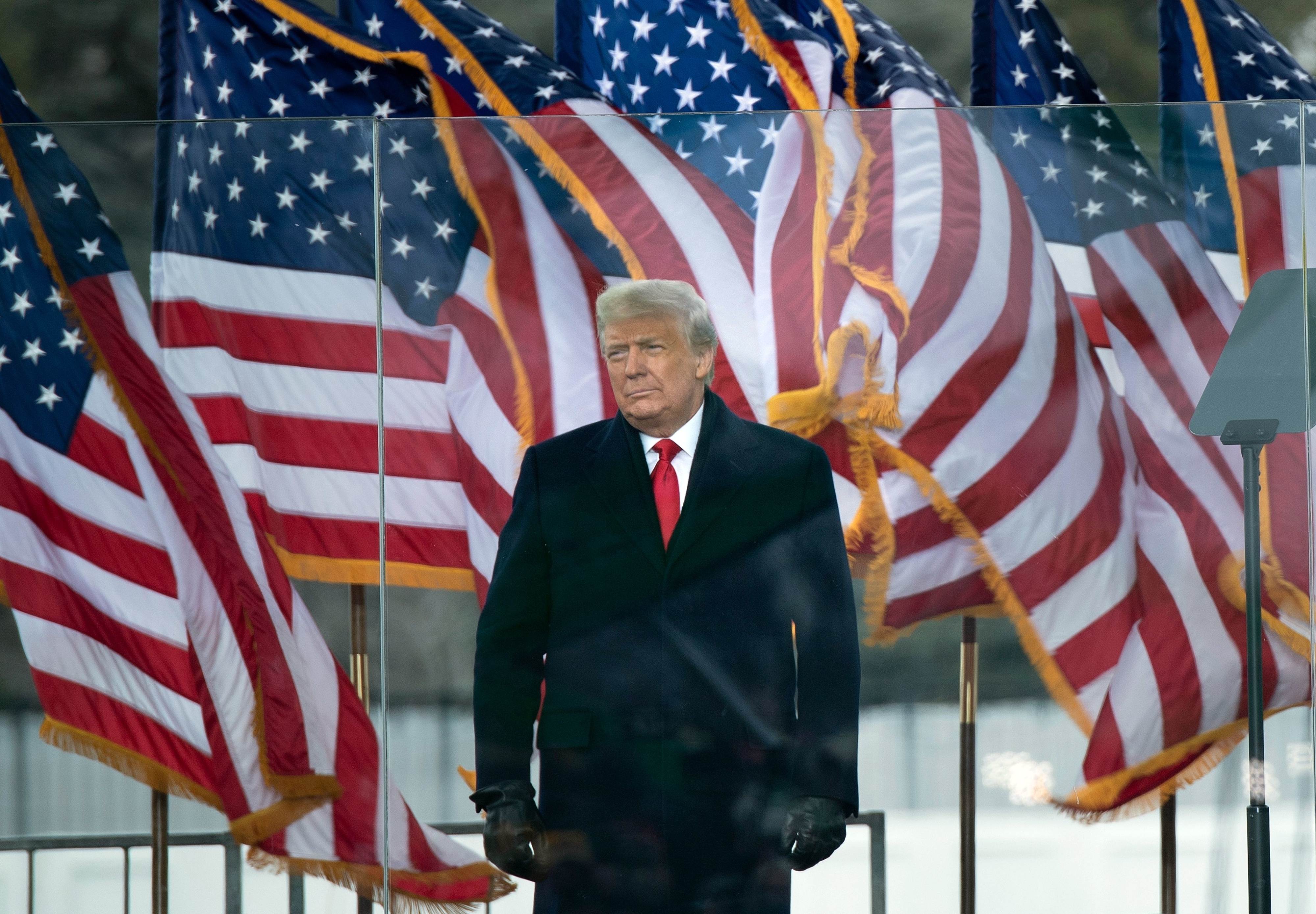 Person in a suit stands outdoors with American flags in the background