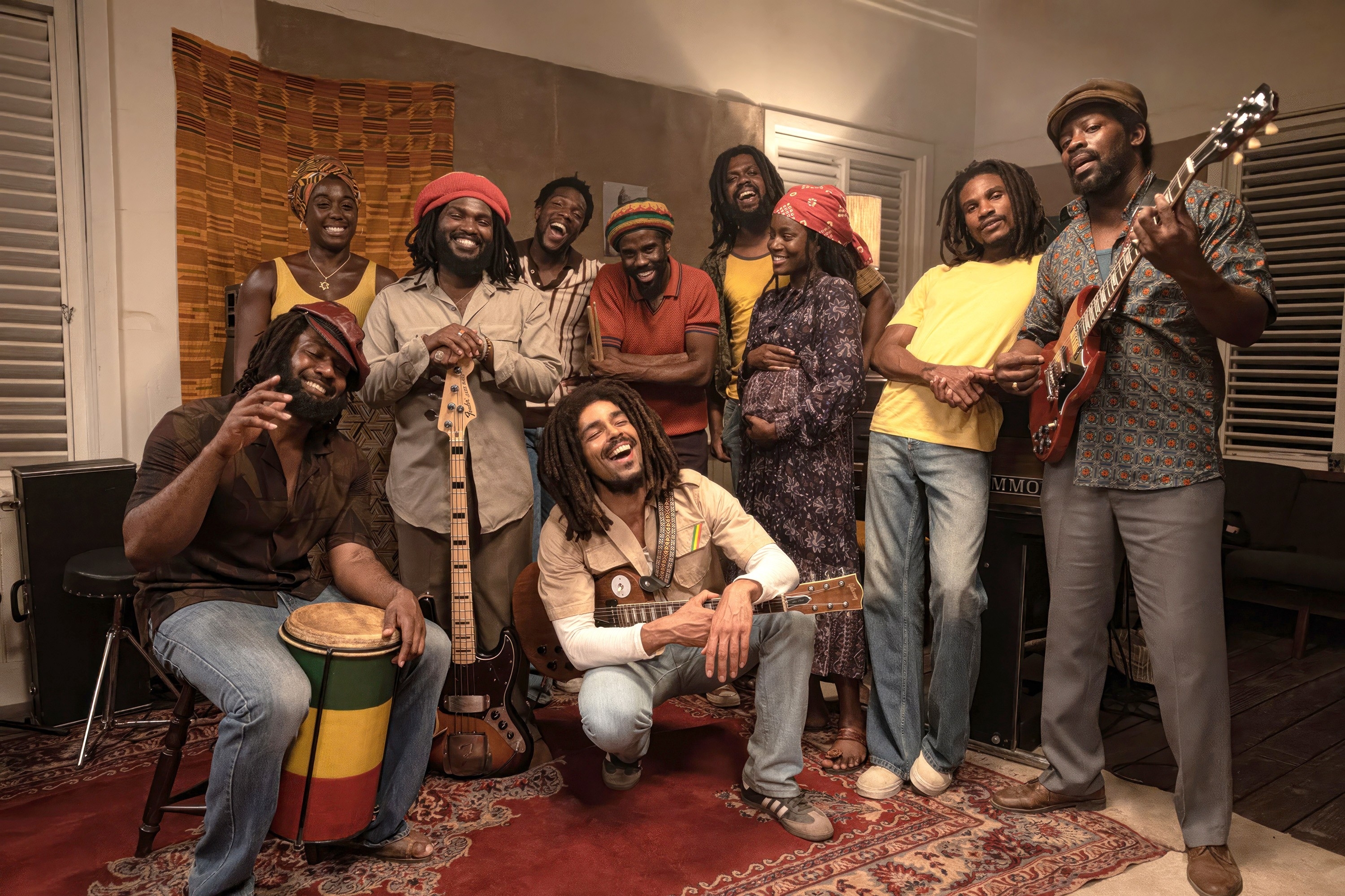 A group of musicians in a scene from Bob Marley: One Love is joyfully gathered in a room, with one playing guitar and another with a drum. They are casually dressed and smiling