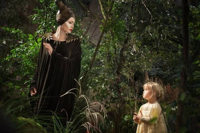 A scene from a fantasy movie with a woman in a dramatic horned costume and a child in a simple dress, standing in a forest setting