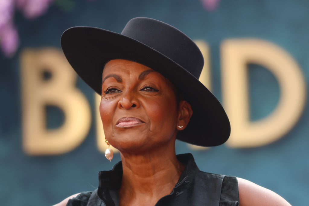 A person in a stylish wide-brimmed hat and sleeveless top stands against a blurred backdrop, appearing confident and elegant