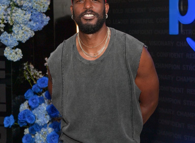 Luke James  wearing a sleeveless shirt and hat poses in front of a backdrop with flowers at a celebrity event