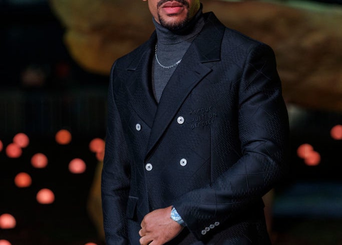 Aaron Pierre in a dark suit poses outdoors at a nighttime event with decorative lights in the background