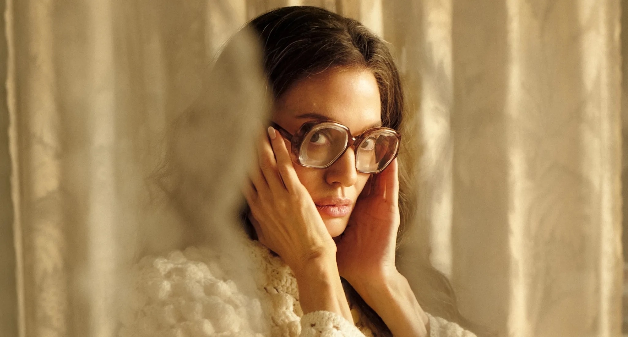 Person wearing glasses, peering through sheer curtains in a contemplative pose