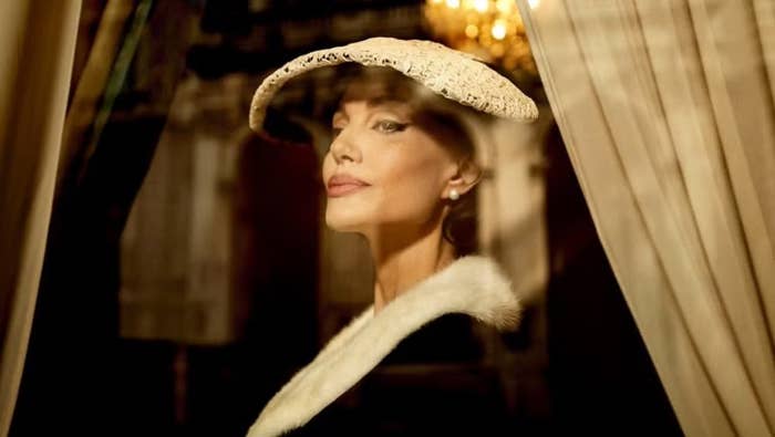 Woman in elegant attire with a lace hat and fur-trimmed coat, looking through a window with draped curtains, creating a classic, glamorous vibe