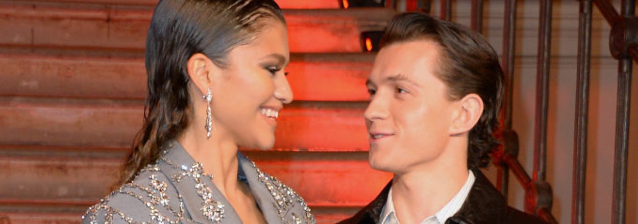 Zendaya and Tom Holland smile at each other. One wears a jeweled blazer, the other a leather jacket. The setting appears elegant and formal