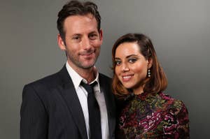 Jeff Baena in a suit and tie stands next to Aubrey Plaza wearing a floral-patterned high-neck dress, both smiling at the camera against a neutral background