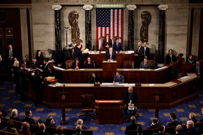 U.S. Congress session in progress with officials seated and discussing. An American flag is displayed above the speaker&#x27;s podium