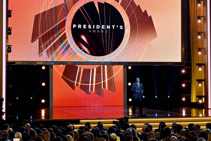 A person stands on stage at an awards ceremony, with a large screen displaying &quot;President&#x27;s Award&quot; in the background
