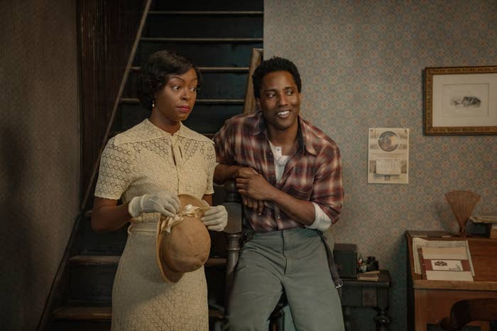 Two people in vintage-style clothing stand indoors, one holding a hat. The setting suggests a historical period