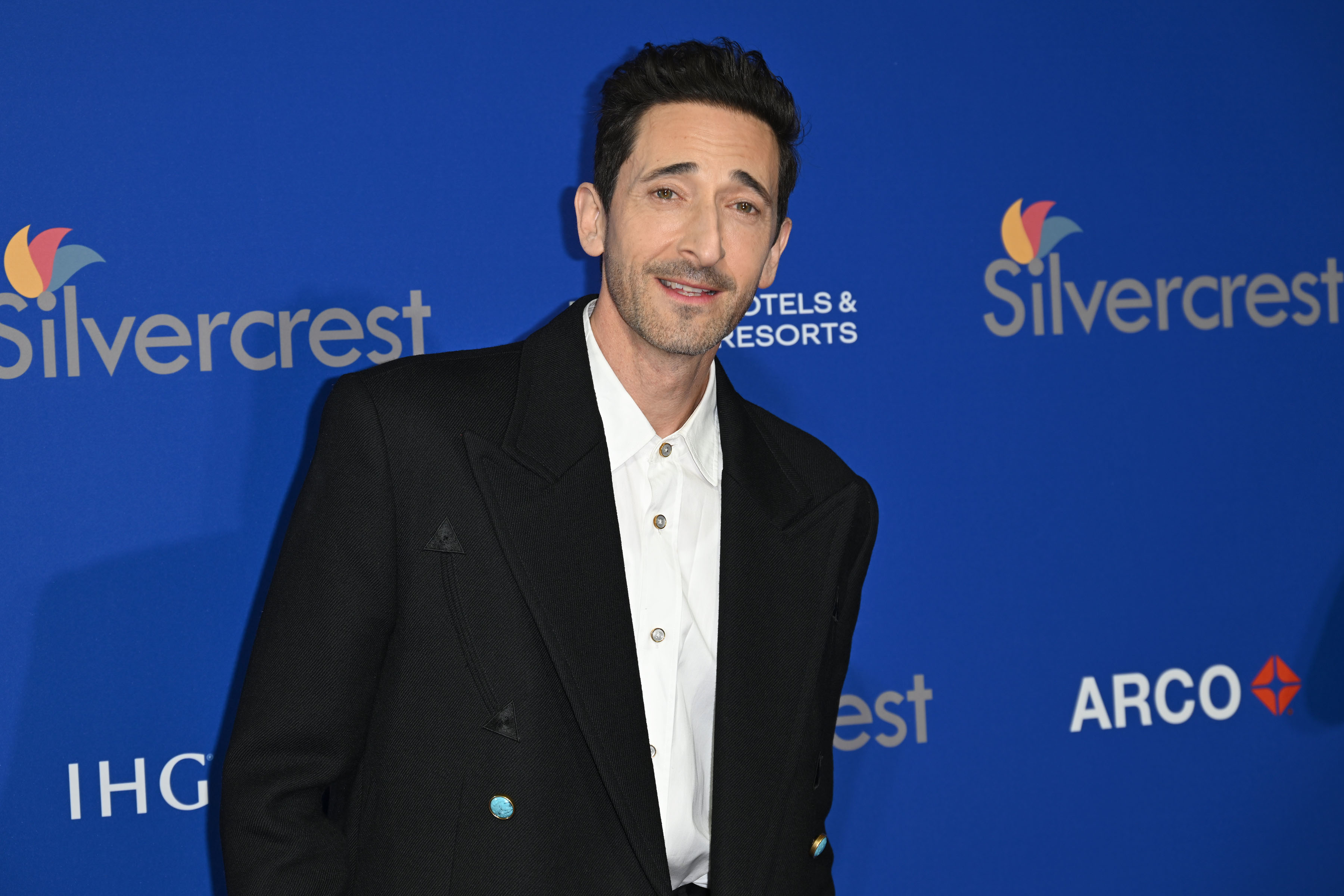 Adrien Brody in a formal black jacket with a white shirt standing at an event with promotional logos in the background