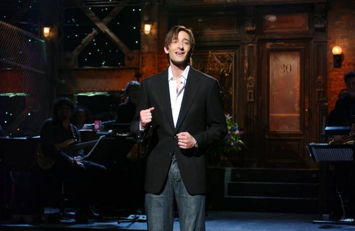 Adrien Brody wearing a black blazer, white shirt, and jeans, likely performing or making an announcement. Background includes musicians