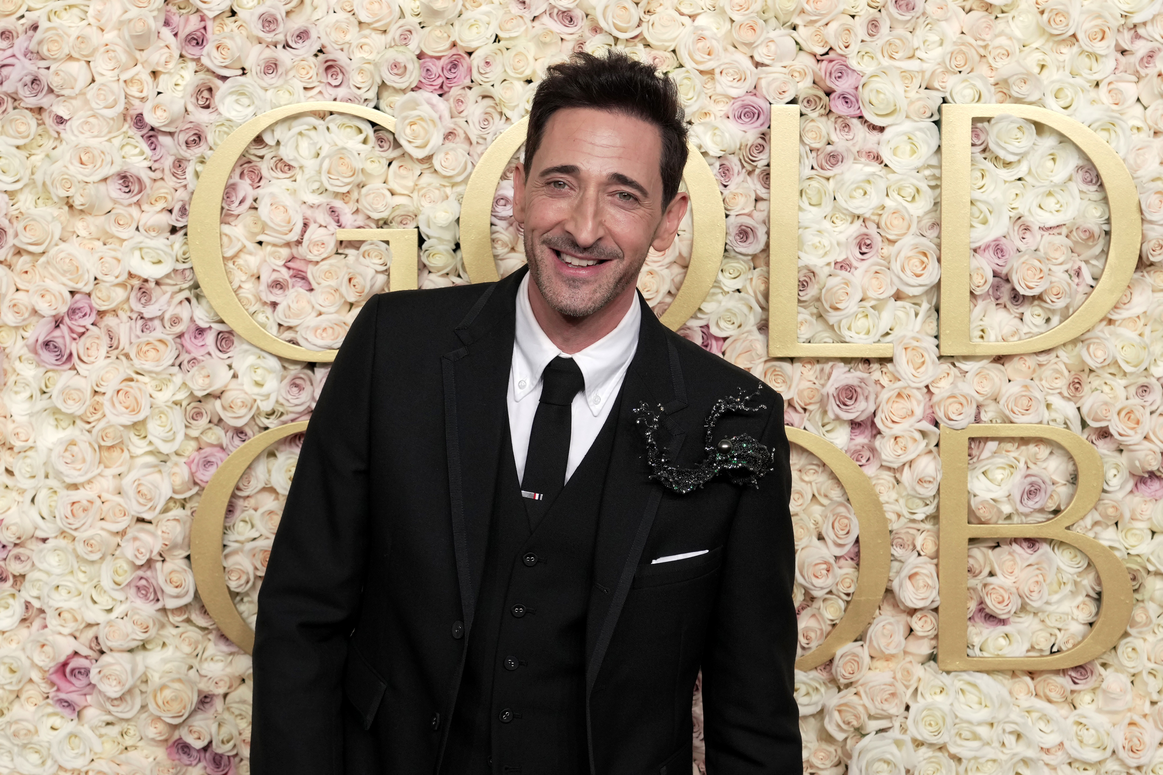 Adrien Brody in a black suit with a floral backdrop smiles at a formal event