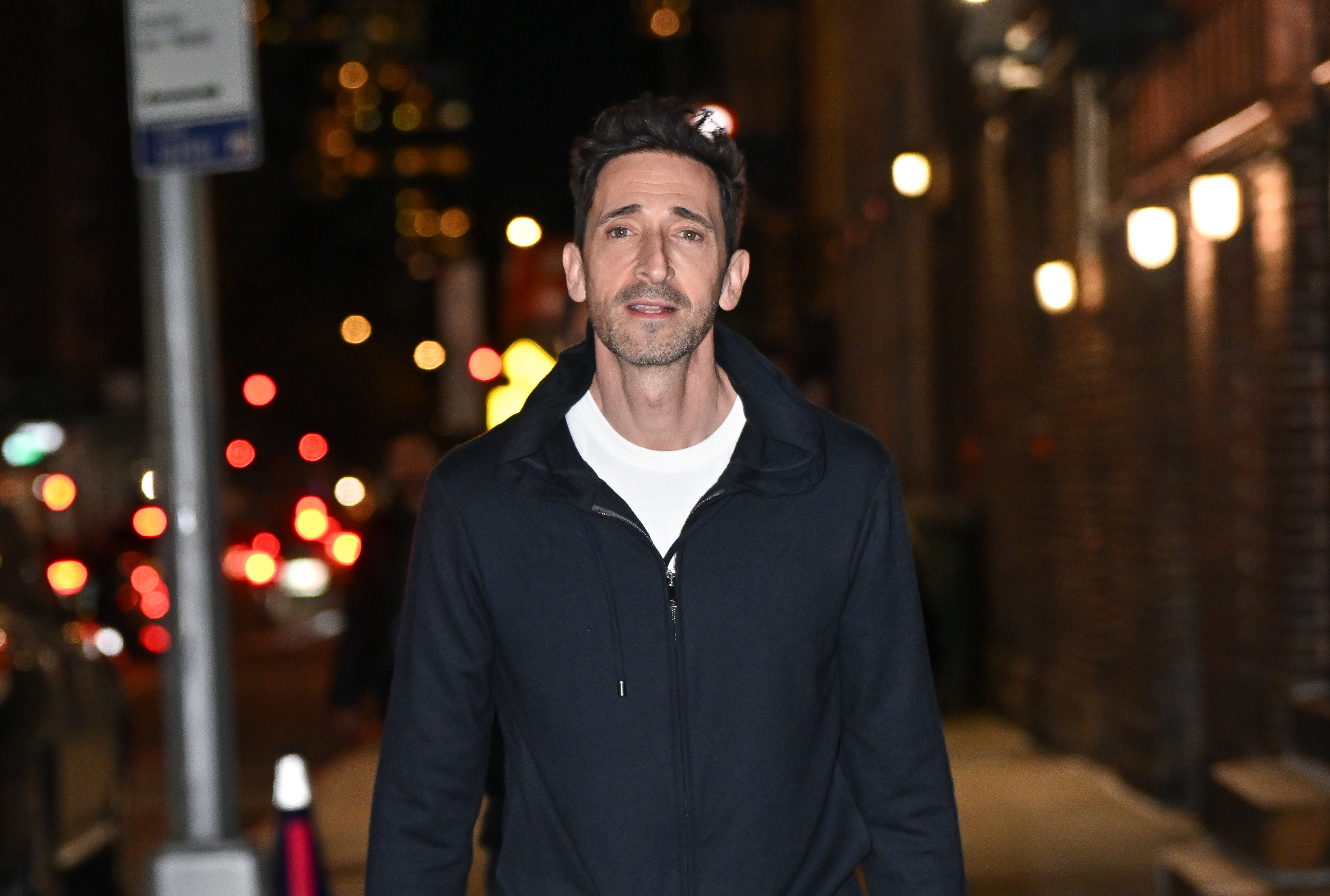 Adrien Brody is walking down an urban street at night, wearing a casual jacket over a t-shirt. The background has blurred city lights and buildings