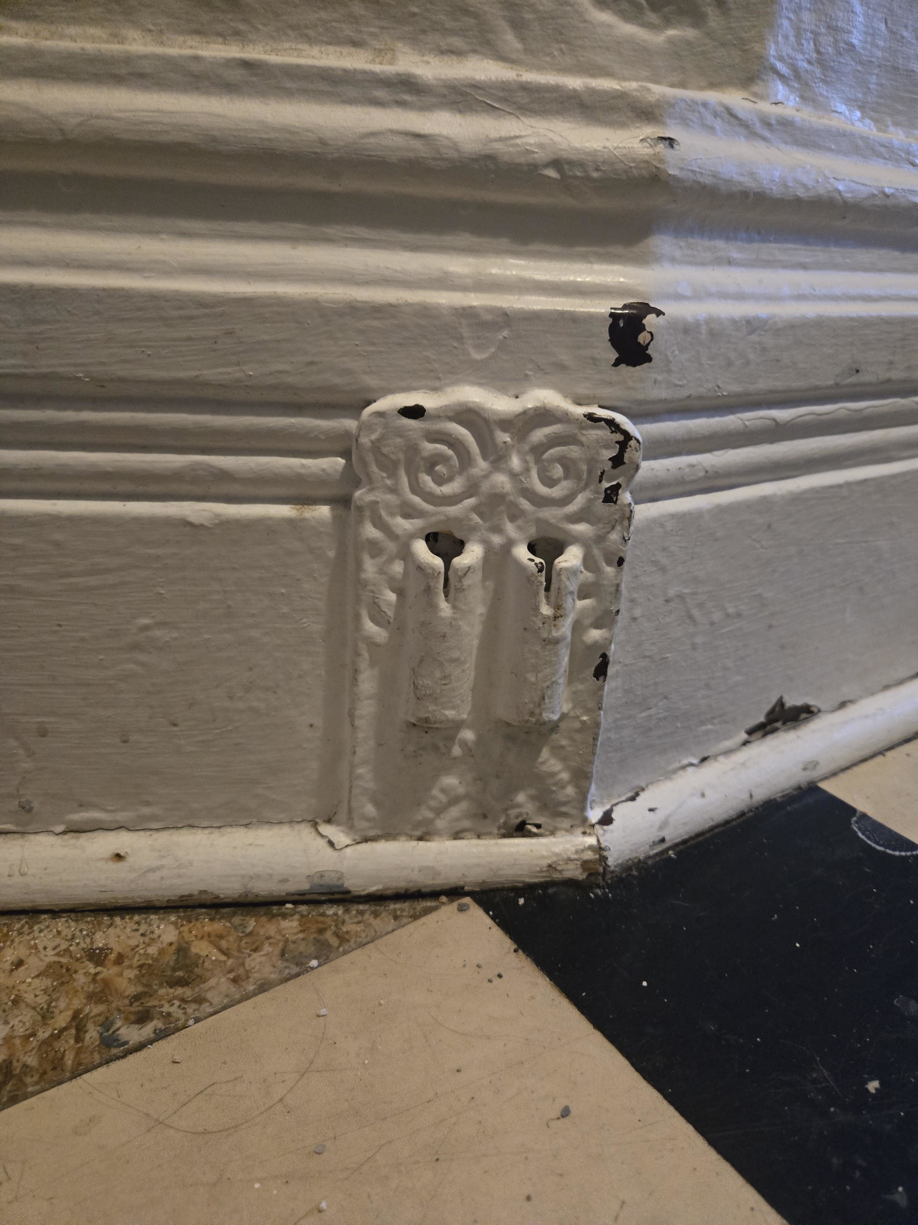Decorative vintage wall detail with two ornate hooks, set above a black and white checkered floor. The paint is slightly chipped