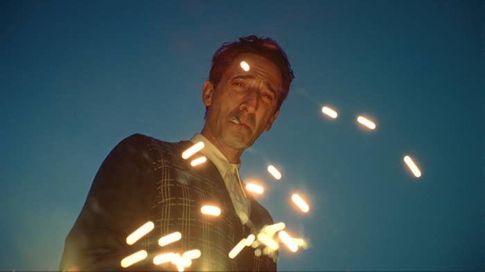 Adrien Brody wearing a plaid suit jacket stands against a sky backdrop with sparklers creating arcs of light around them