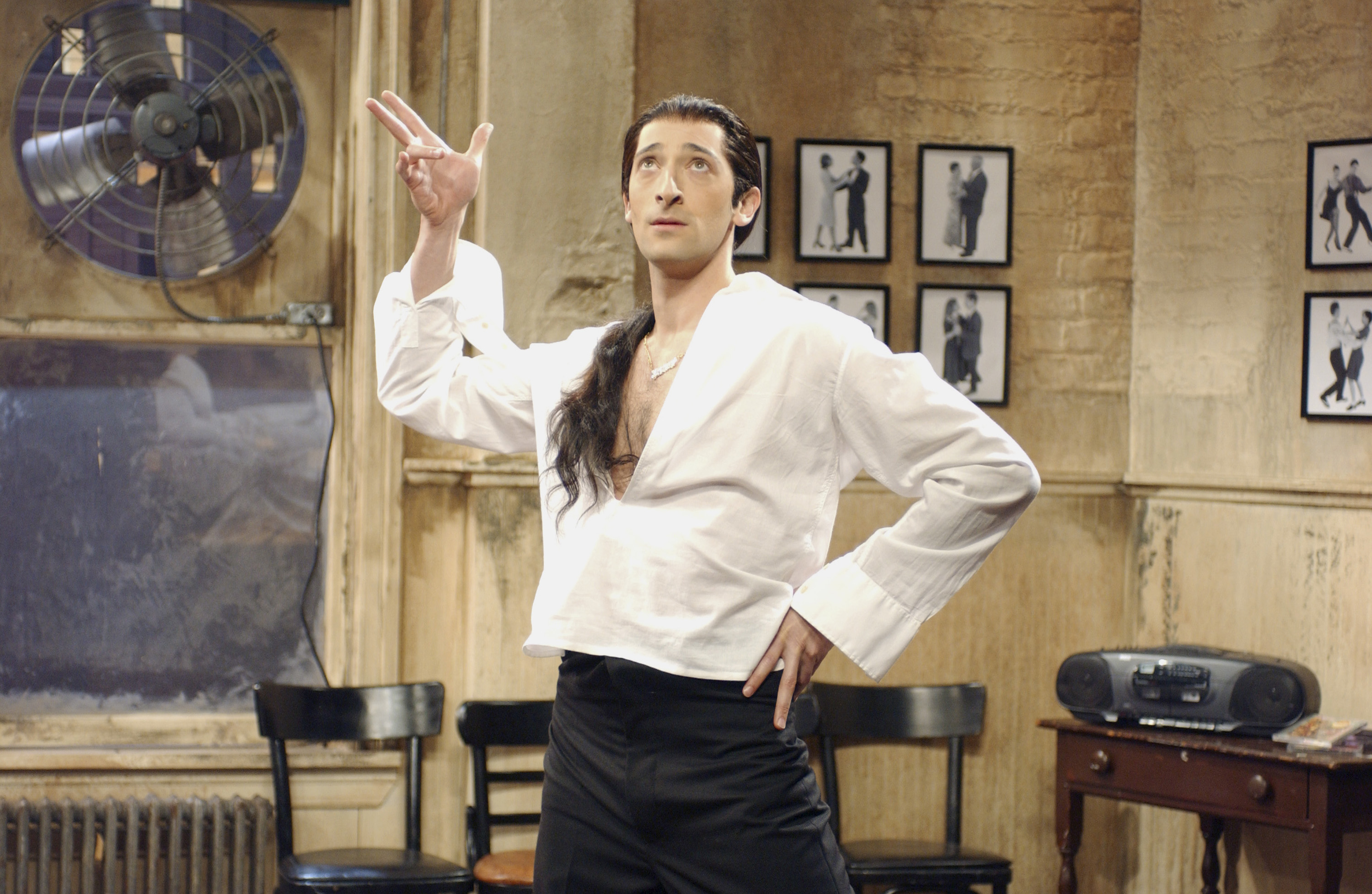 Adrien Brody in a white shirt and black pants strikes a dramatic pose in a room with framed photos and wooden chairs