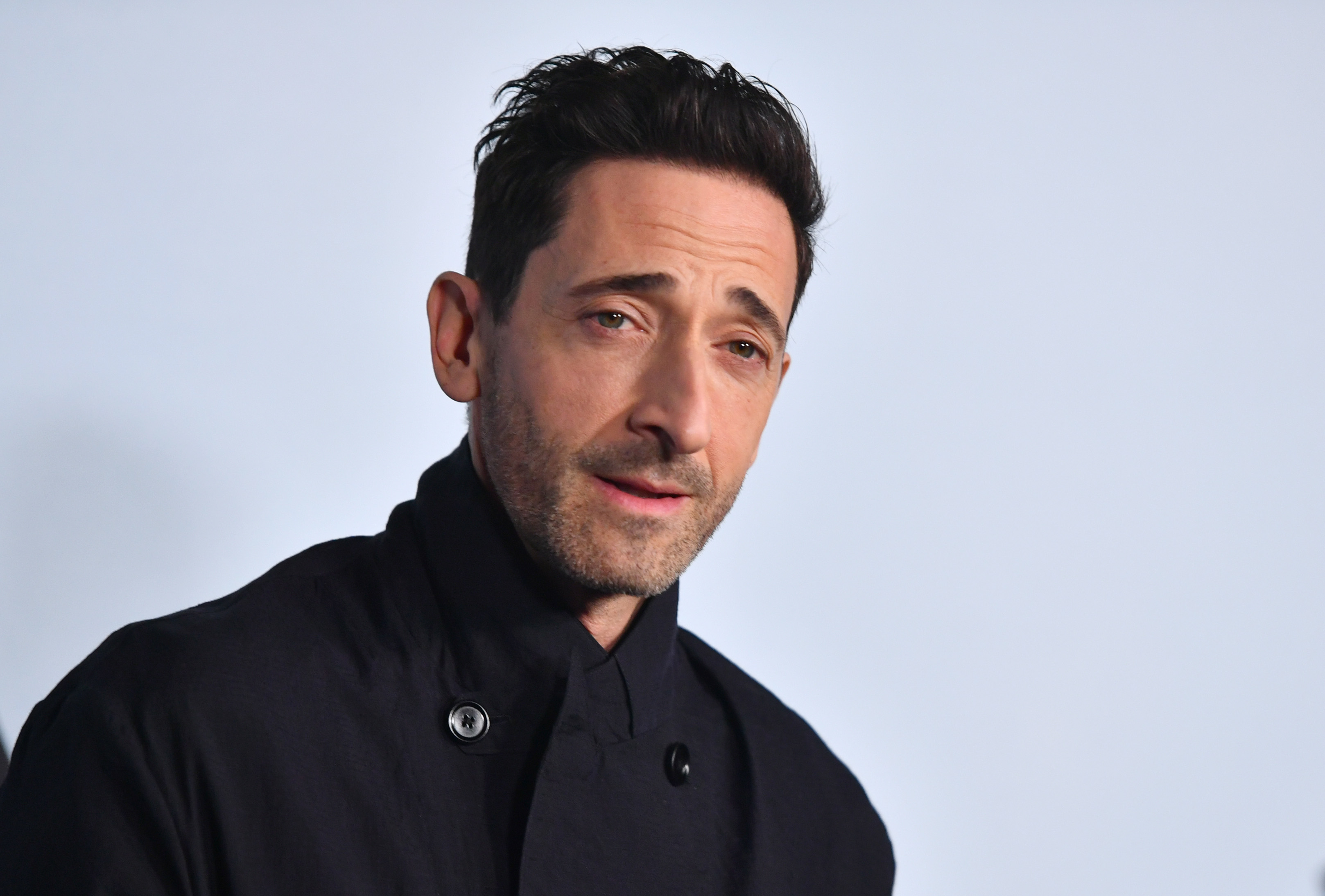 Adrien Brody in a buttoned black coat against a plain background