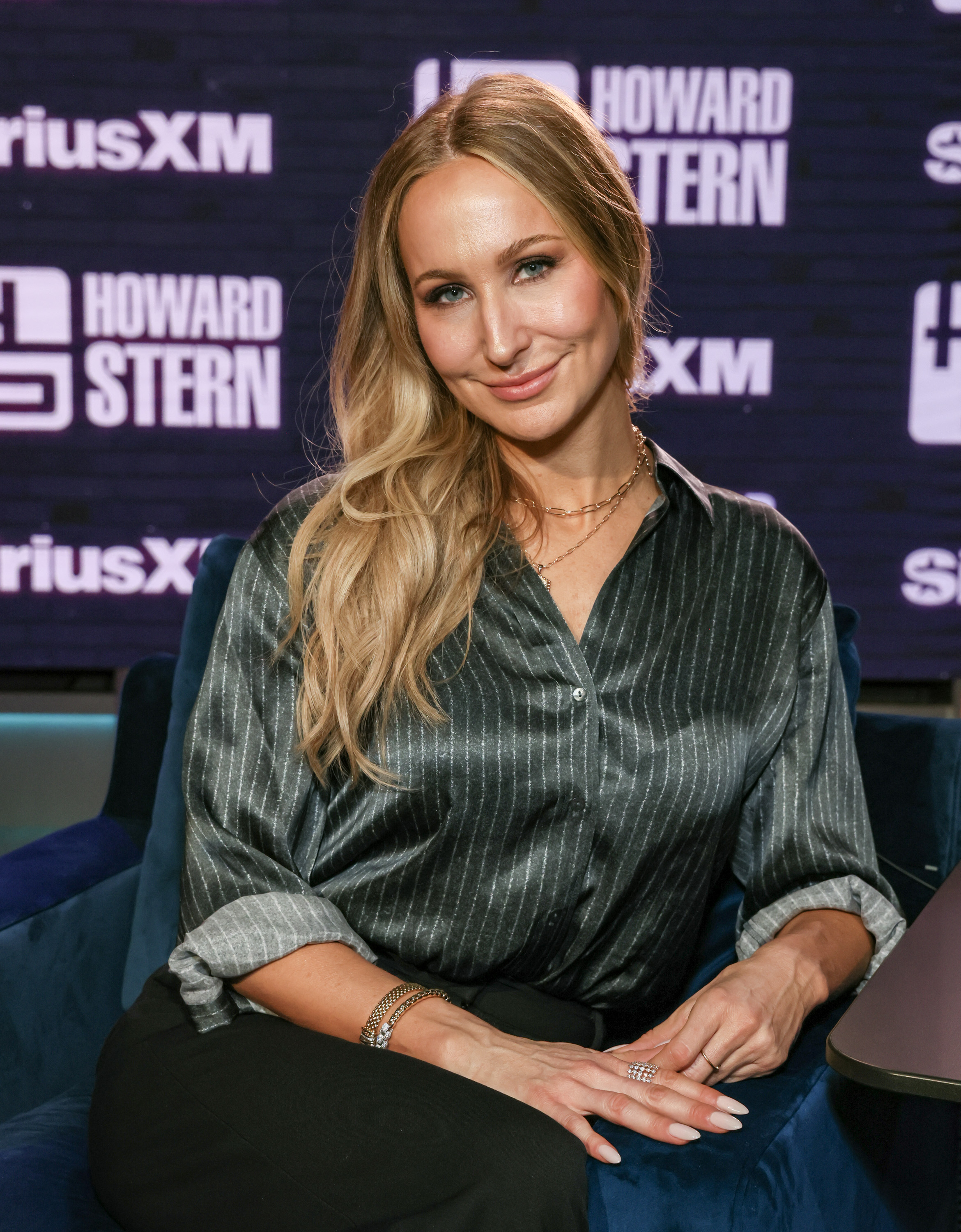 Person in a pinstriped blouse sits smiling in a studio setting with Howard Stern show logos in the background