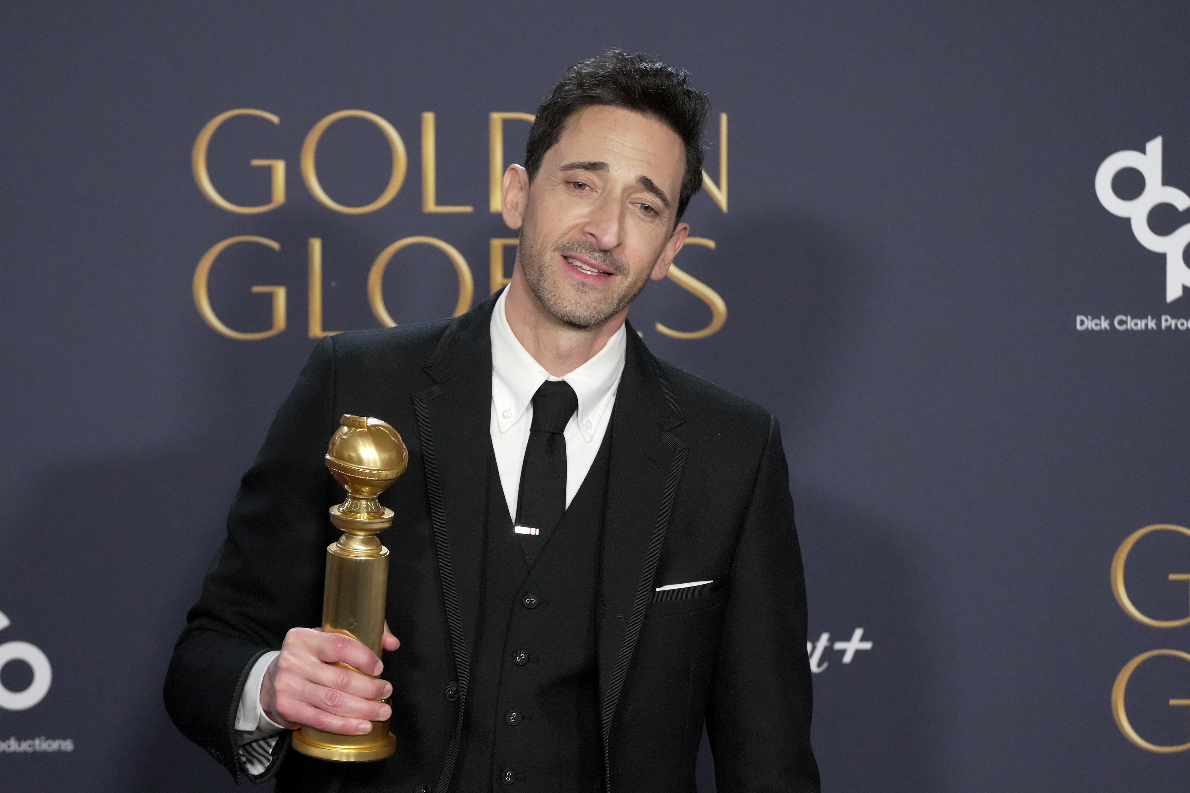 Adrien Brody holding a Golden Globe award, wearing a dark suit and tie, standing in front of a backdrop with Golden Globes branding