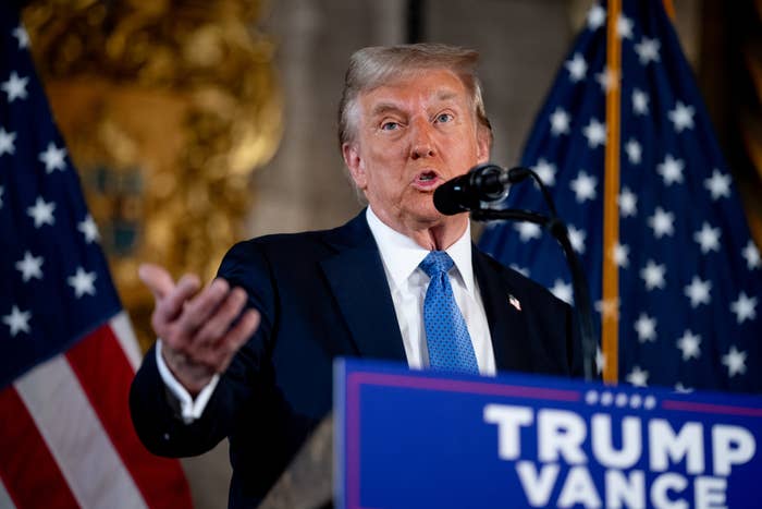 Person speaking at a podium with a microphone, surrounded by American flags. A sign with &quot;TRUMP&quot; and &quot;PENCE&quot; is visible in the foreground