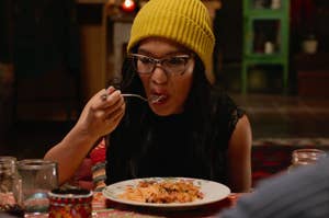 Ali Wong wearing glasses and a yellow beanie is eating spaghetti with a fork at a table