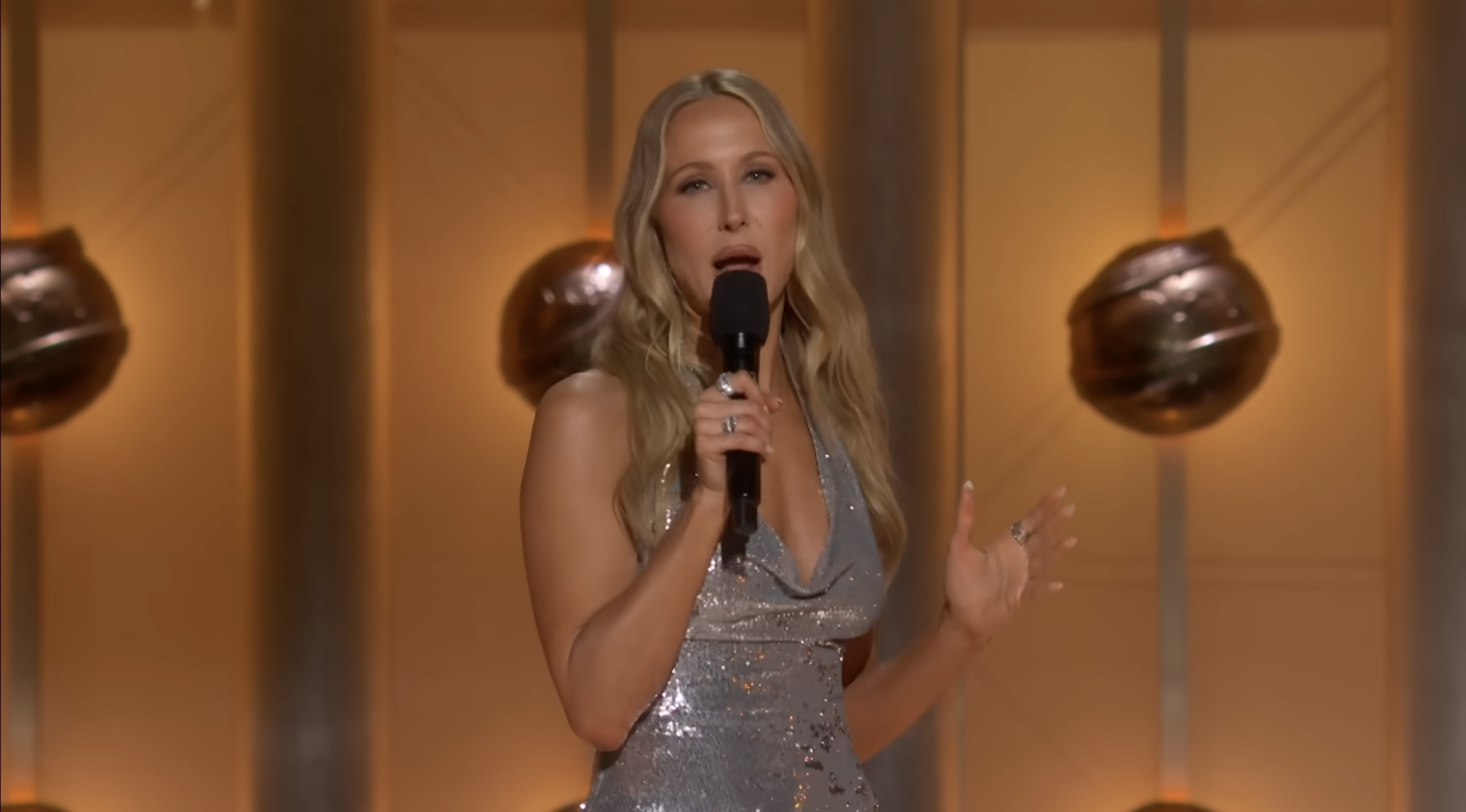 Person presents on stage in a sparkly draped dress, holding a microphone against a backdrop with spherical lights