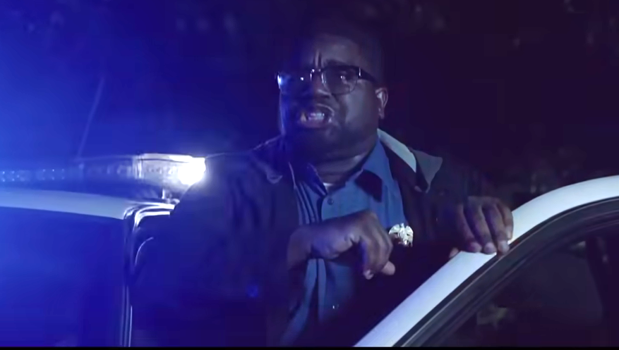 A man wearing glasses and holding a badge sits inside a police car at night, illuminated by flashing lights, appearing to shout urgently