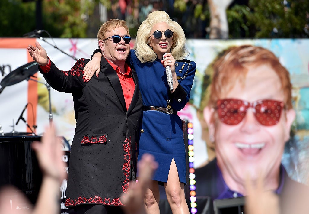  one in a decorated suit with sunglasses, the other in a stylish dress with a microphone, both smiling and embracing