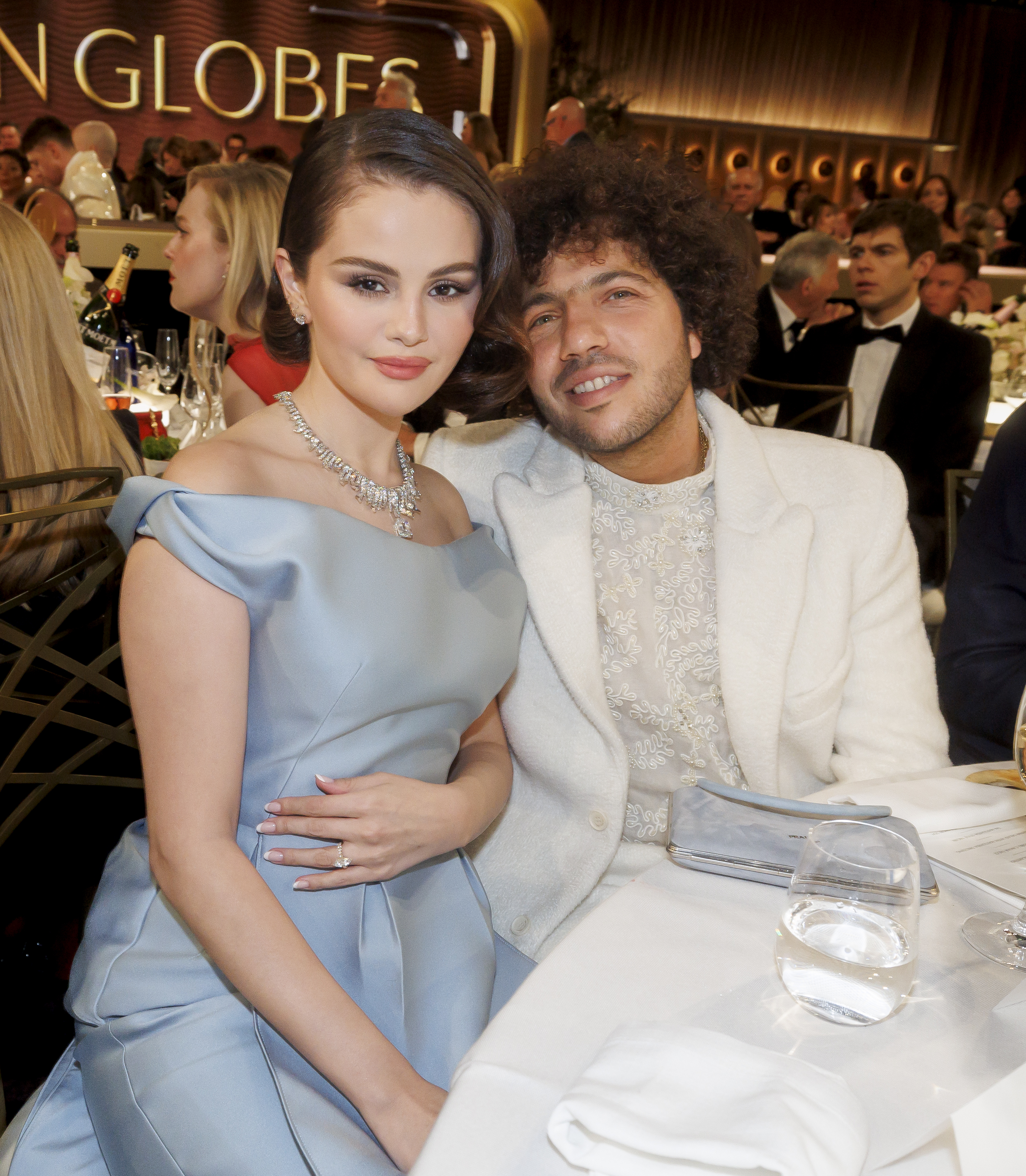 Two people seated at a formal event, one in an elegant off-shoulder gown and the other in a suit, posing for a photo at a table setting