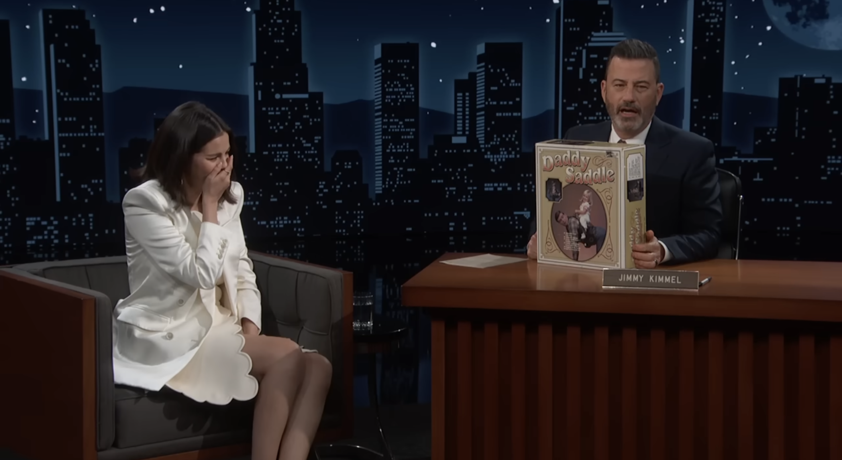 Talk show host on set showing a comedic gift box to a guest in a white outfit, who is laughing