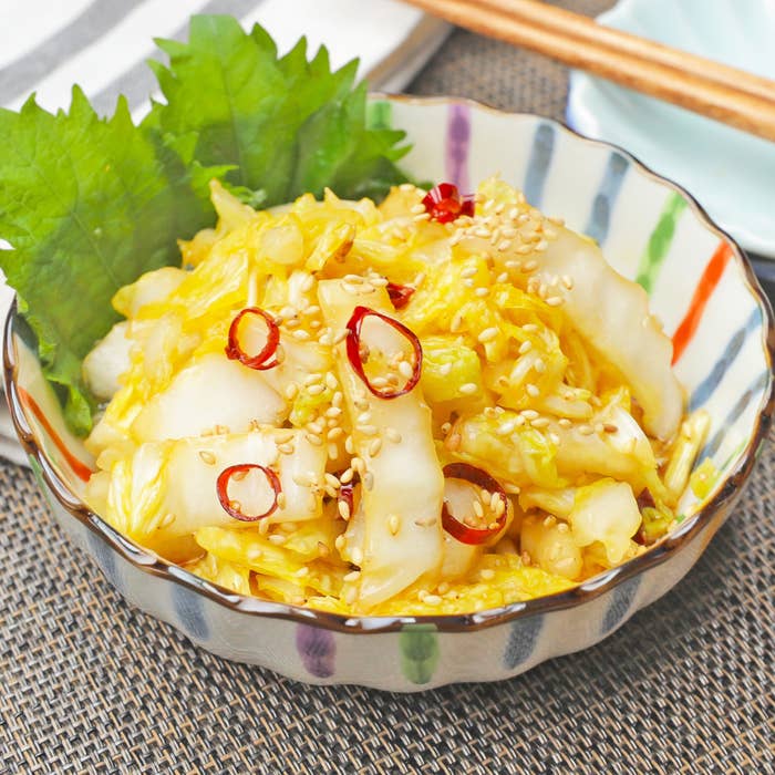 白菜と唐辛子の漬物がボウルに盛られている。ゴマがトッピングされており、バックに葉が添えられている。