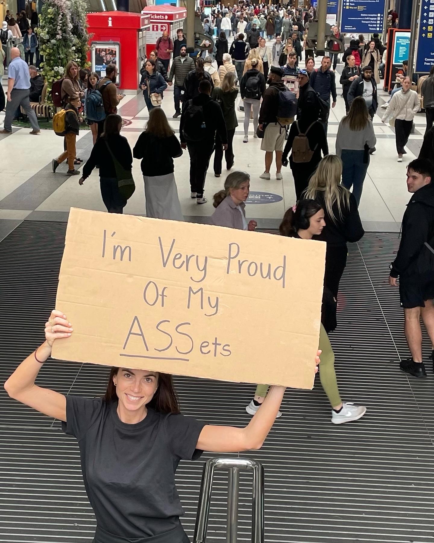 Person holds a cardboard sign reading &quot;I&#x27;m Very Proud Of My ASSets&quot; in a crowded public space